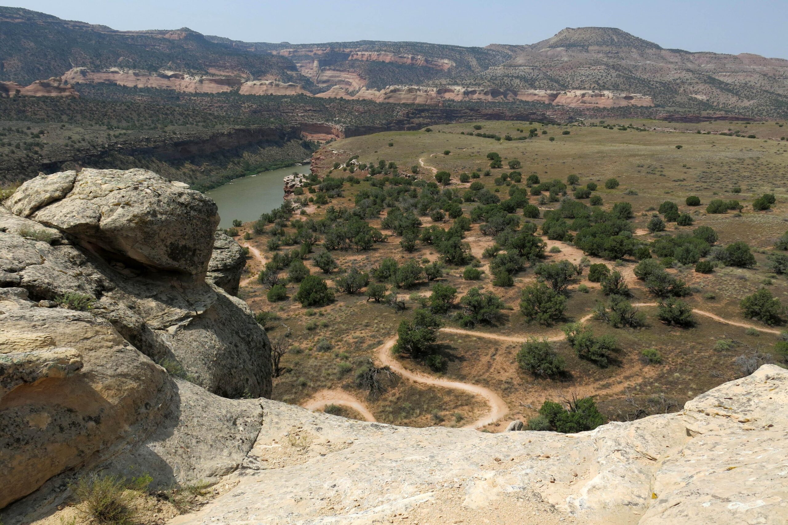 A panoramic view of a rugged landscape featuring a winding river surrounded by rocky cliffs and rolling hills. The foreground showcases large boulders and shrubs, while the lower area displays a sandy path meandering through patches of green vegetation. The scene is bathed in bright sunlight, highlighting the natural textures and colors of the terrain. Mary's Loop / Horsethief Bench mountain bike trail.