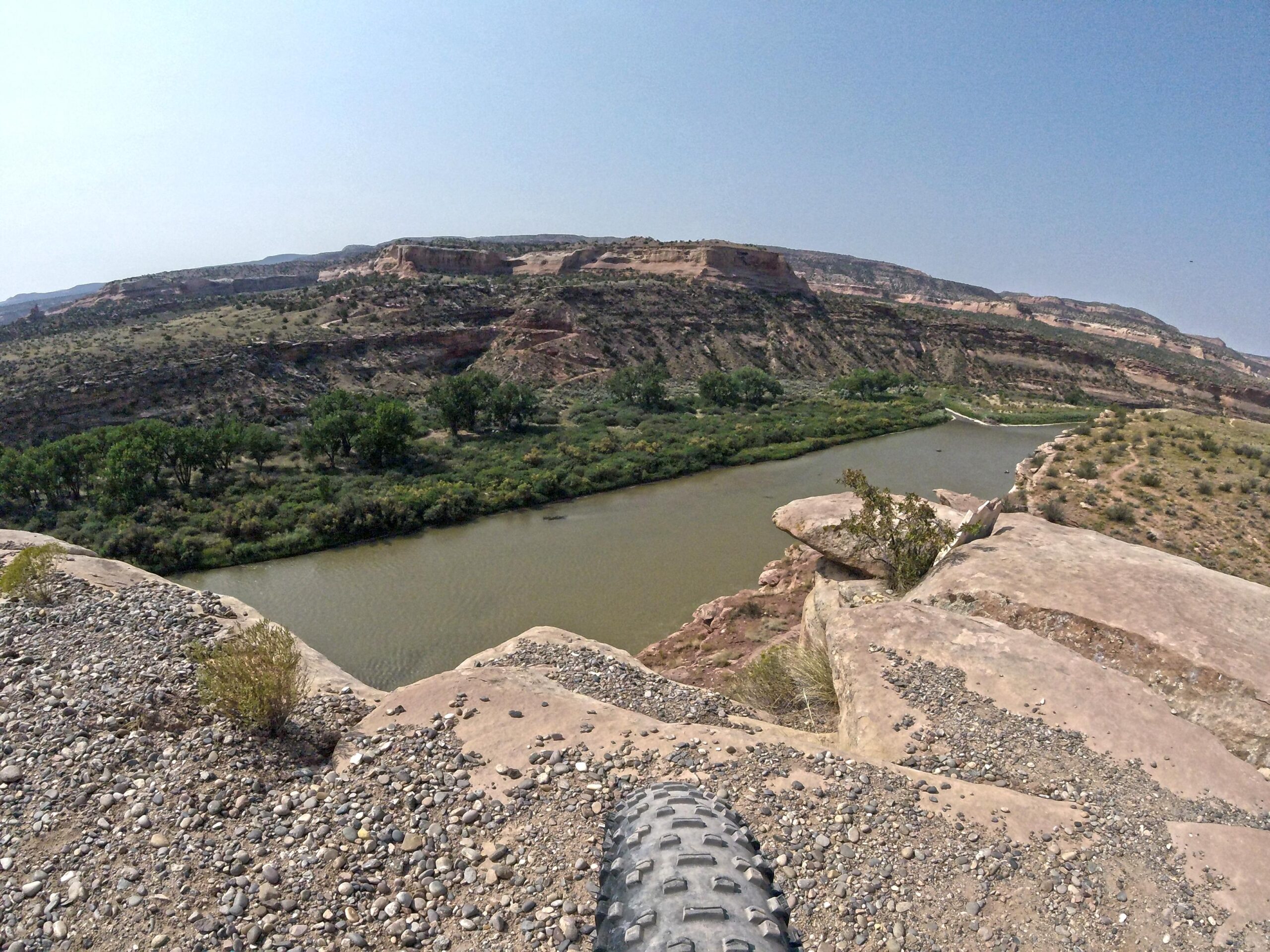 A panoramic view of a river winding through a rocky landscape, with steep cliffs on either side. The foreground includes gravel and rocks, showcasing a mountain bike tire, while the background features lush green trees along the riverbank and rocky hills under a clear blue sky. Mary's Loop / Horsethief Bench mountain bike trail.