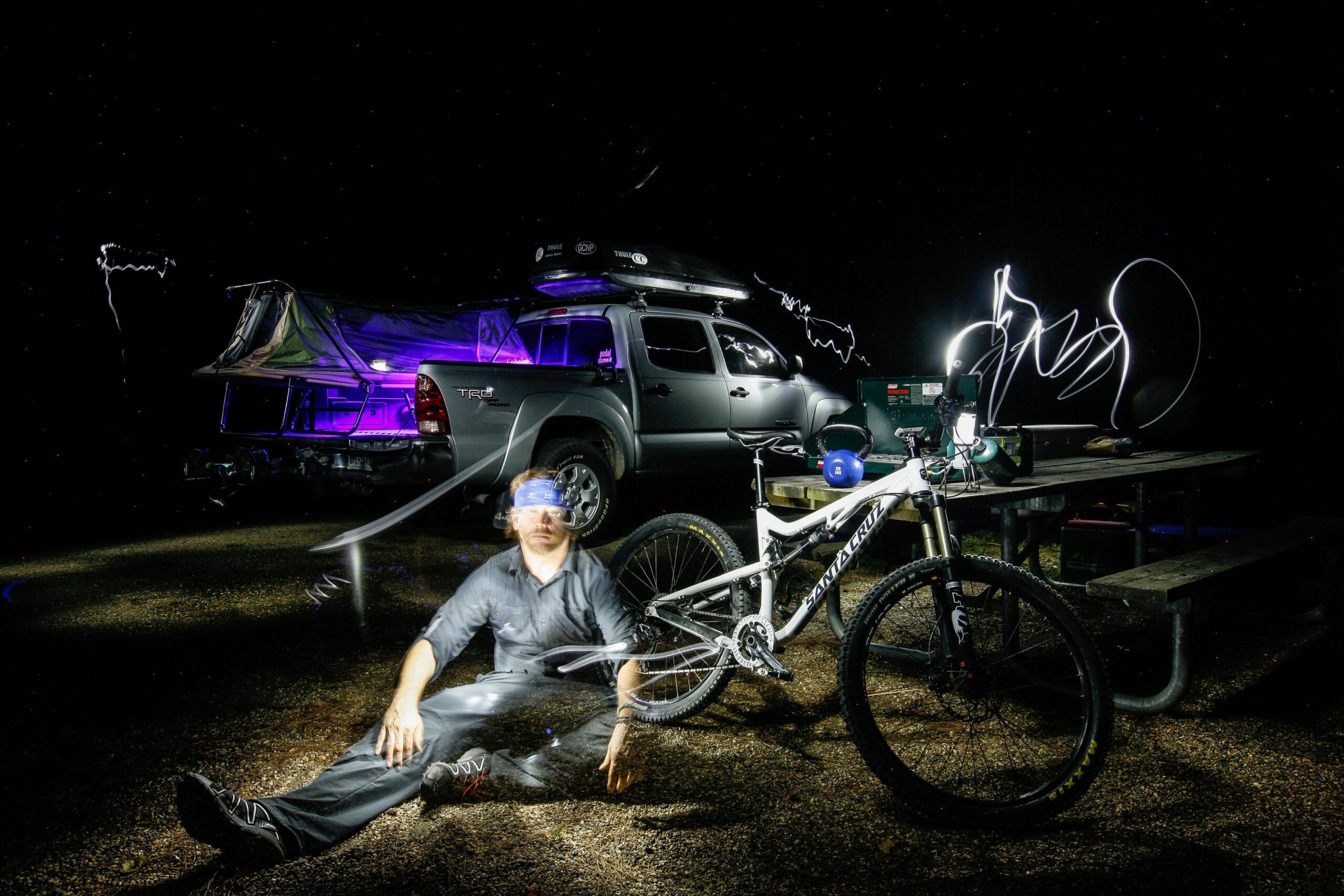Santa Cruz 5010: A person wearing a blindfold is sitting on the ground next to a mountain bike, with a pickup truck and a campsite setting illuminated by colorful lights in the background. Light trails can be seen in the air, creating an artistic effect against the night sky. A picnic table is visible nearby, adding to the outdoor atmosphere.