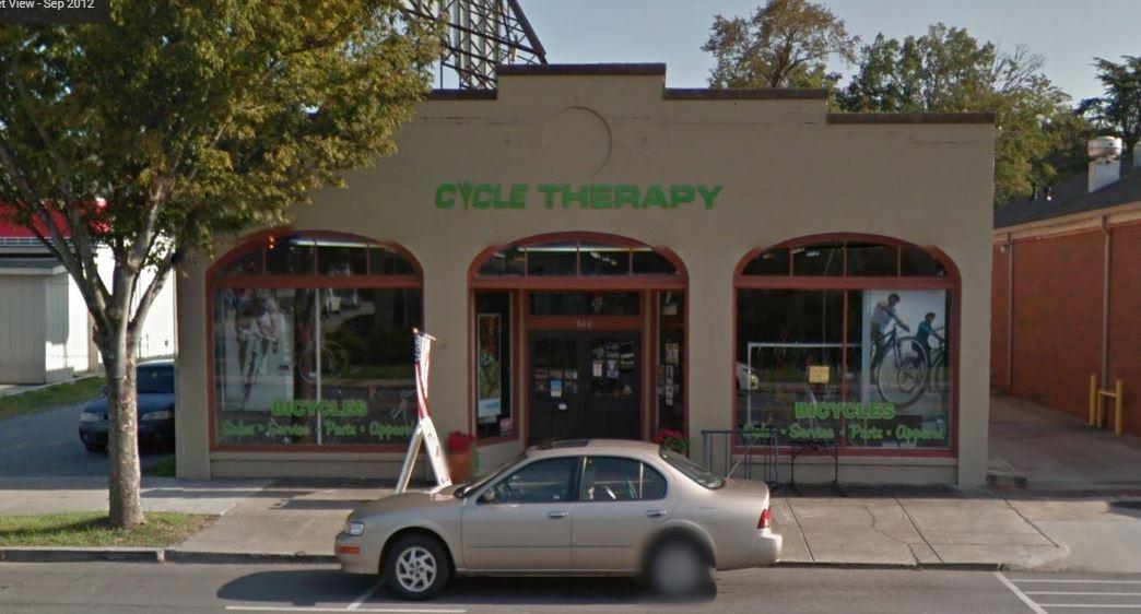 A storefront for "Cycle Therapy," featuring large windows displaying bicycles and accessories. The business name is written in green above the entrance. A light-colored car is parked in front, with trees providing shade on either side of the building.