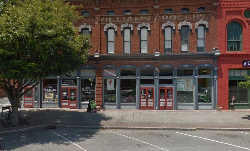 A historic brick building featuring storefronts, including a hair shop and a pharmacy, with decorative trim around the windows and a large tree in front. The building has a faded sign reading "WILLIAM