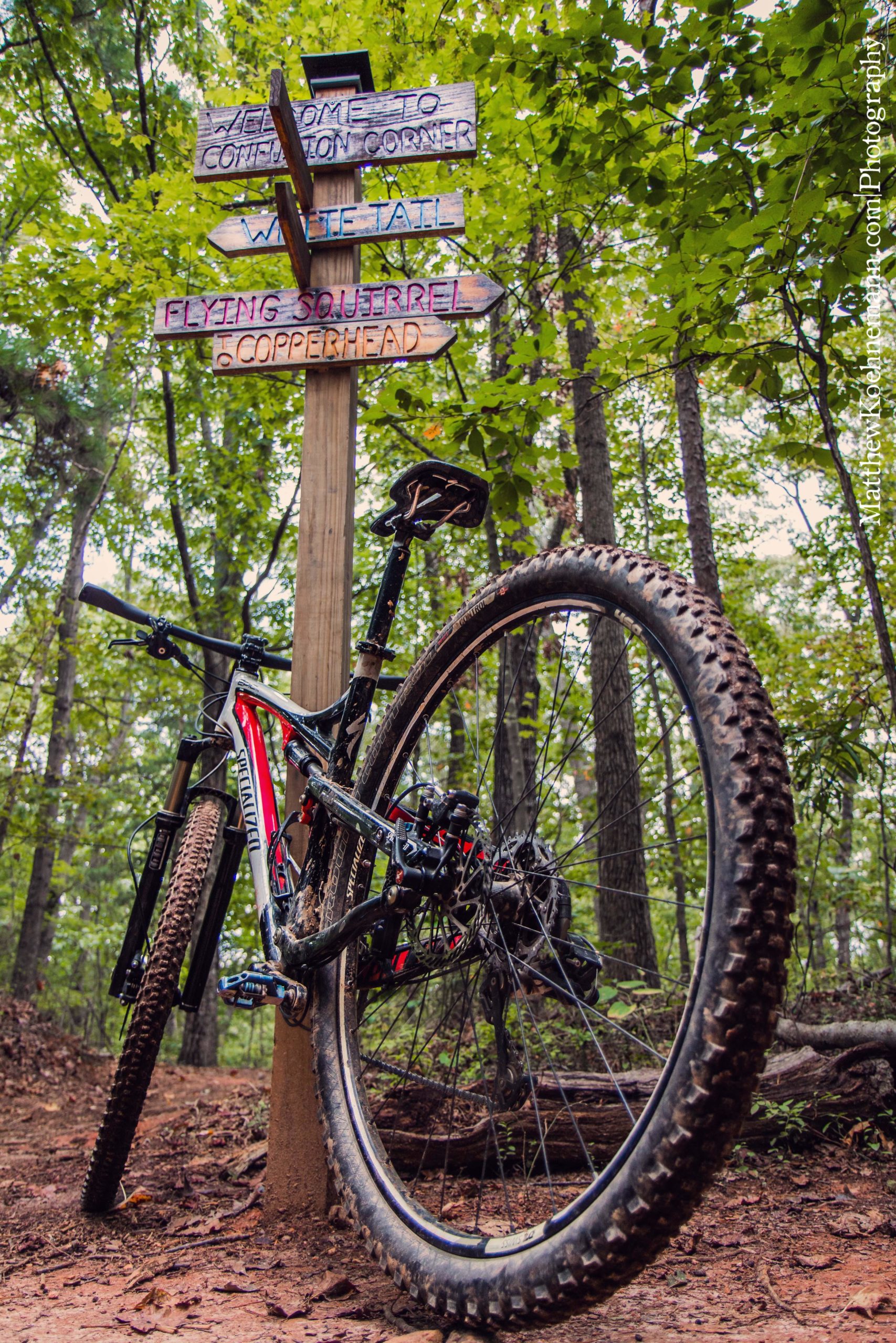 A mountain bike leaning against a wooden signpost with multiple arrows indicating different trail names. The area is surrounded by lush green trees, showcasing a serene forest trail. The bike is partially covered in dirt, suggesting recent use on a muddy path. Chicopee Woods mountain bike trail.