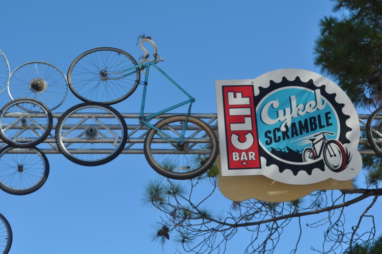 A blue bicycle is creatively displayed among several bicycle wheels on a metal structure, with a sign featuring the text "CLIF BAR Cykel Scramble." The background is a clear blue sky.