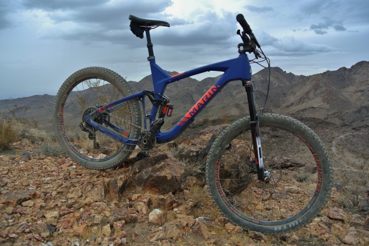 A blue mountain bike leaning against a rocky outcrop, set against a backdrop of rugged mountains and cloudy skies. The bike features knobby tires and a sleek design, highlighting its suitability for off-road trails.
