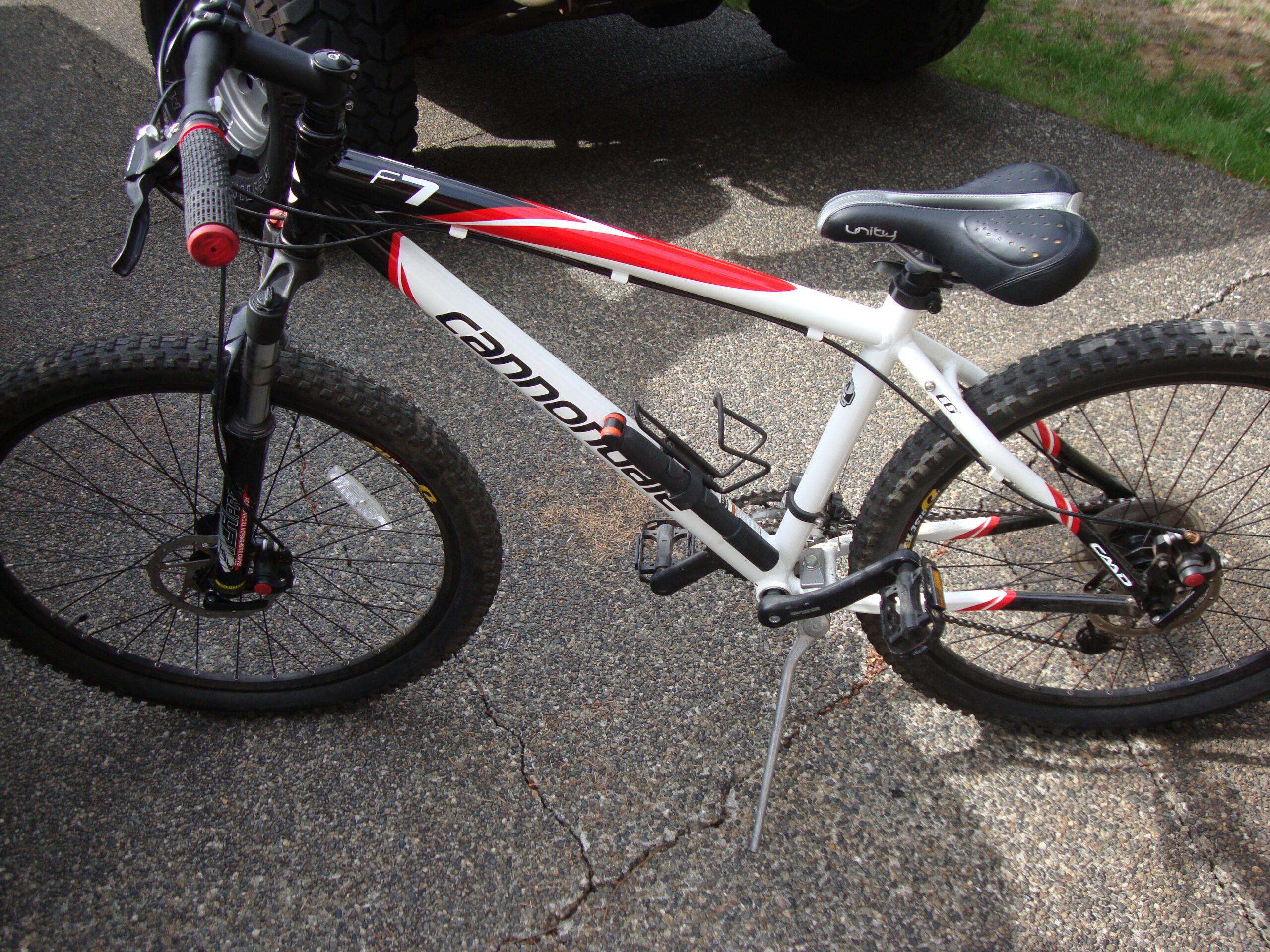 Cannondale F7: A Cannondale F7 mountain bike, primarily white with red and black accents, featuring thick tires, a sturdy frame, and a black saddle. The bike is parked on a textured asphalt surface, with a kickstand deployed.