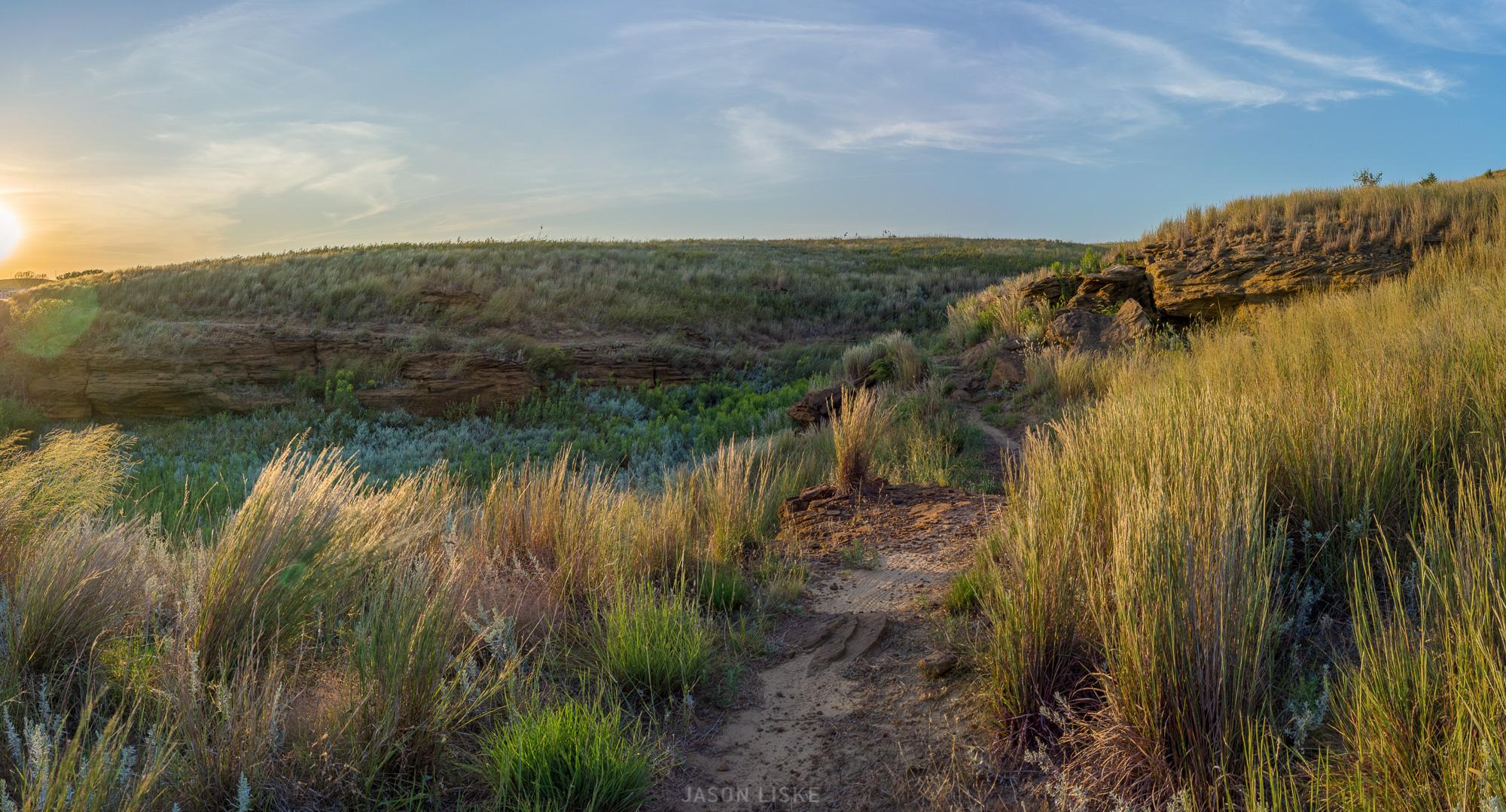 Panoramic view of a grassy landscape with tall grasses and a rocky cliff. The sun is setting on the horizon, casting a warm golden light over the scene, while wispy clouds are visible in the sky. A sandy path winds through the vegetation, inviting exploration of this natural setting. Switchgrass mountain bike trail.