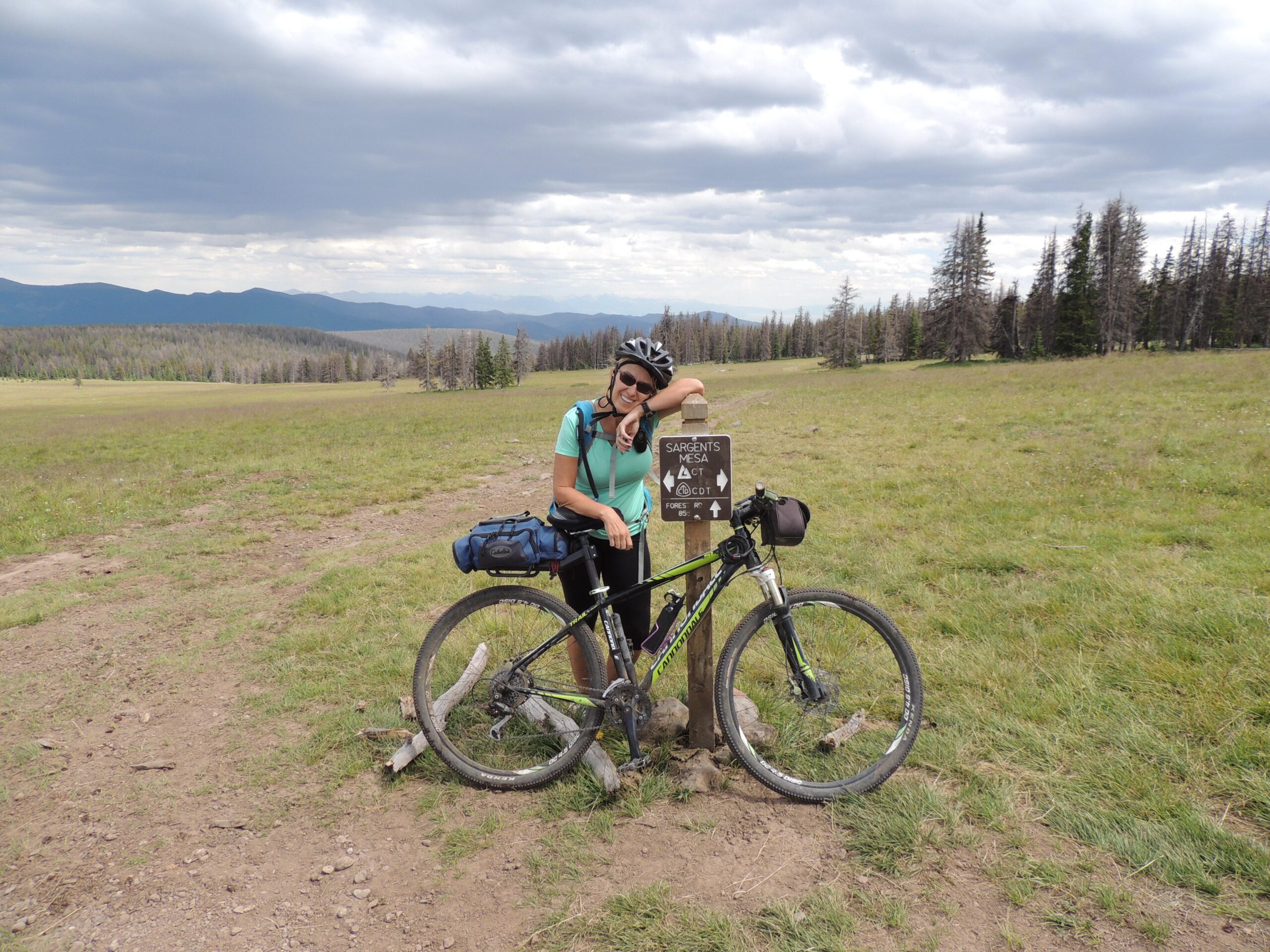 Cannondale Trail SL4: A woman with a bicycle stands beside a trail sign marked "Sargents Mesa." She is wearing a helmet and a teal shirt, smiling as she leans against the sign. The background features a green meadow with distant mountains and cloudy skies.