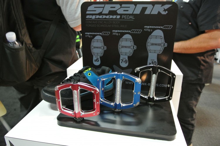 Alt text: "Three Spank Spoon pedals displayed on a table, featuring red, blue, and black colors. A black sneaker with blue accents is positioned next to the pedals. A sign behind the pedals lists their specifications, including weight and size recommendations."
