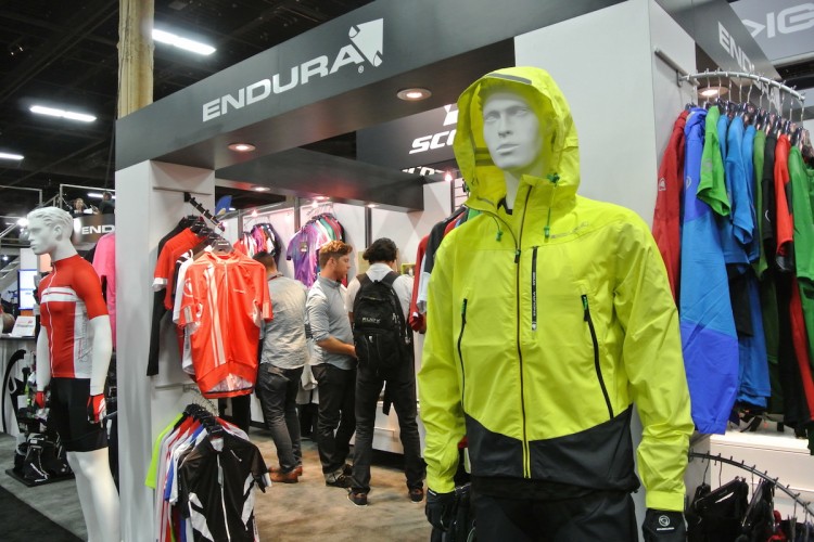 A vibrant trade show display featuring cycling apparel from Endura. In the foreground, a mannequin dressed in a bright yellow jacket stands beside hanging red and black cycling jerseys. In the background, attendees browse various colorful cycling outfits on display.