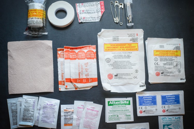 A variety of first aid supplies laid out on a dark surface, including gauze bandages, adhesive tape, non-adherent pads, and assorted medication packets (such as aspirin and diphenhydramine). The arrangement also features easy access bandages, antiseptic towelettes, and insect bite relief products, along with safety pins and a small vial.