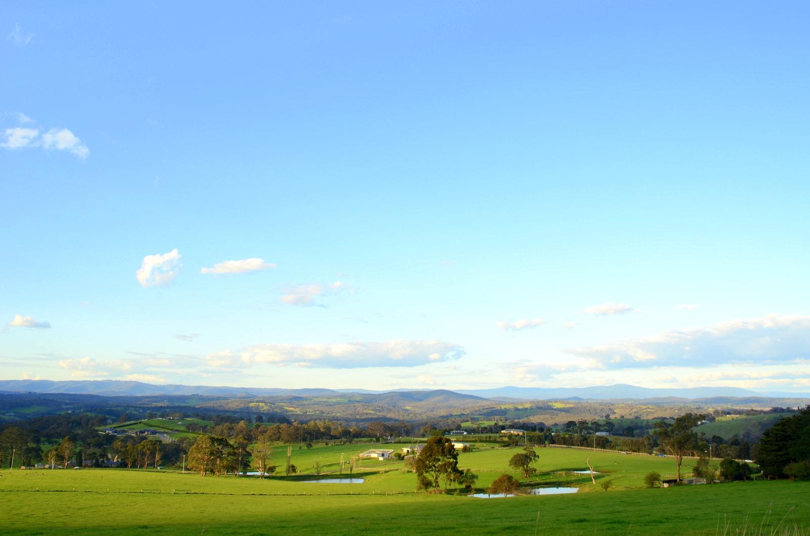 A panoramic view of lush green farmland, rolling hills, and distant mountains under a clear blue sky. Fluffy clouds drift across the horizon, and small ponds reflect the landscape. The scene captures the tranquility of rural life. Smiths Gully mountain bike trail.