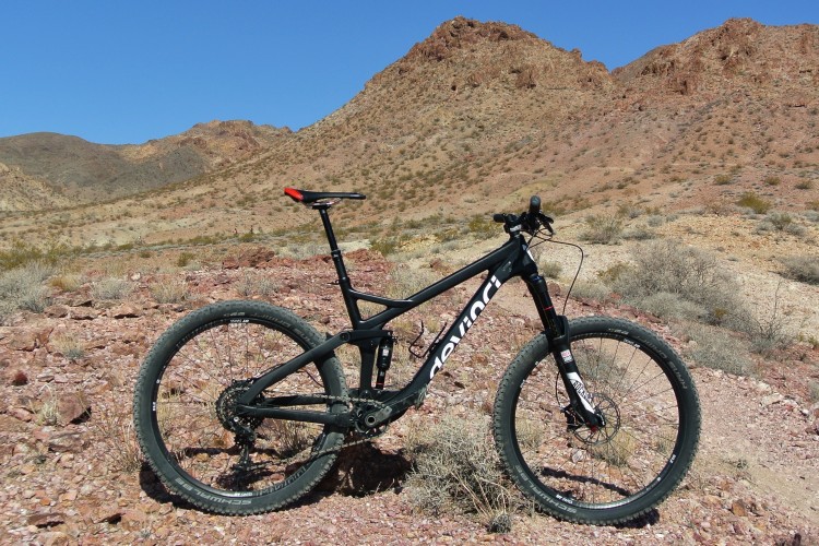 A black mountain bike positioned on rocky terrain with mountains in the background under a clear blue sky.