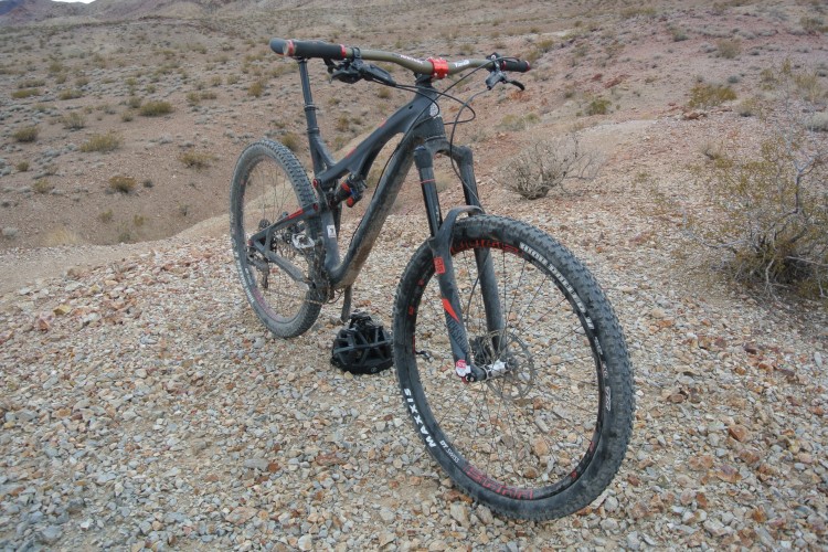 A black mountain bike with red accents rests on a rocky, desert landscape. The bike is positioned at an angle, showcasing its front wheel, handlebars, and rugged tires, which are coated in dirt from off-road use. In the background, sparse vegetation and a mountainous terrain are visible under a cloudy sky.