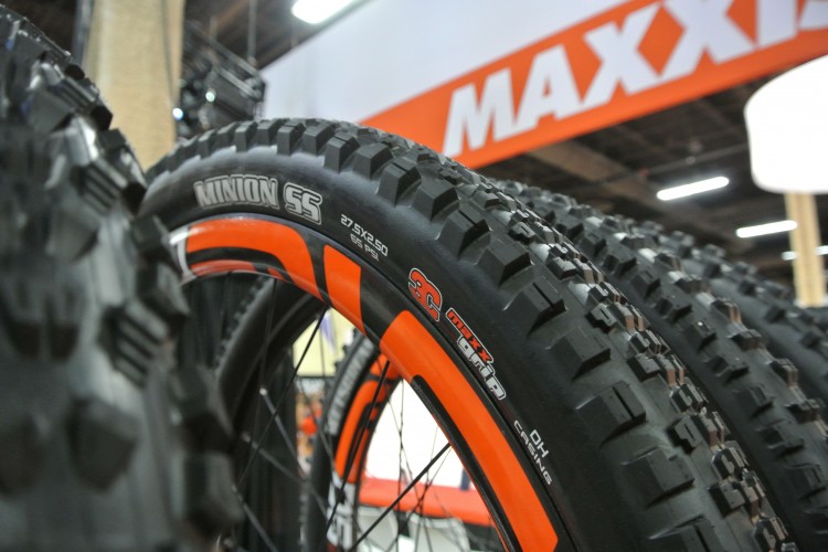 Close-up of Maxxis Minion SS mountain bike tires displaying a textured tread pattern and vibrant orange accents. The image focuses on the tire's sidewall, showcasing details like size and PSI specifications against a blurred background of a display booth.