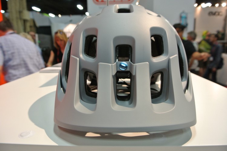 A close-up view of a light gray bicycle helmet placed on a display surface, showcasing its ventilated design and adjustable rear mechanism. The background features blurred figures in a crowded indoor setting, suggesting an event or trade show.