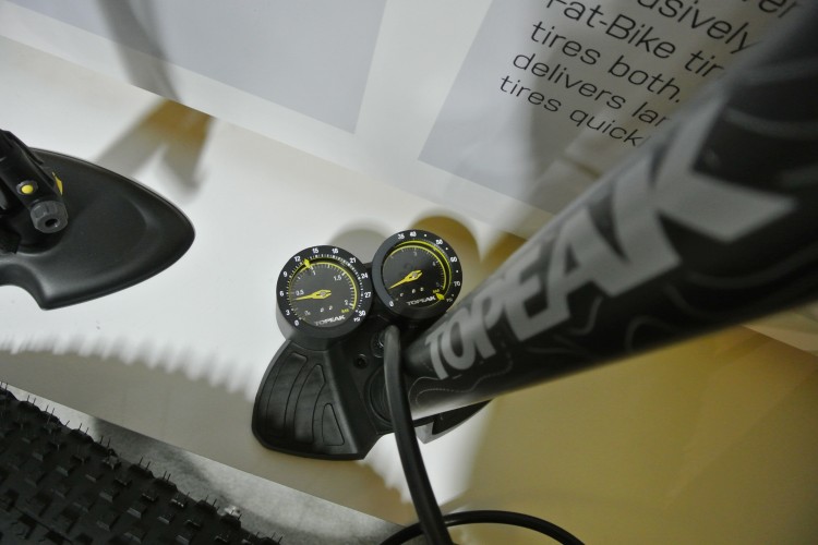 A close-up view of a bike pump with dual pressure gauges, designed for inflating both standard and fat bike tires. The pump features a sturdy base and a hose connected to the gauges, which measure pressure in PSI and Bar. There is a textured background visible, suggesting a workshop or display setting.