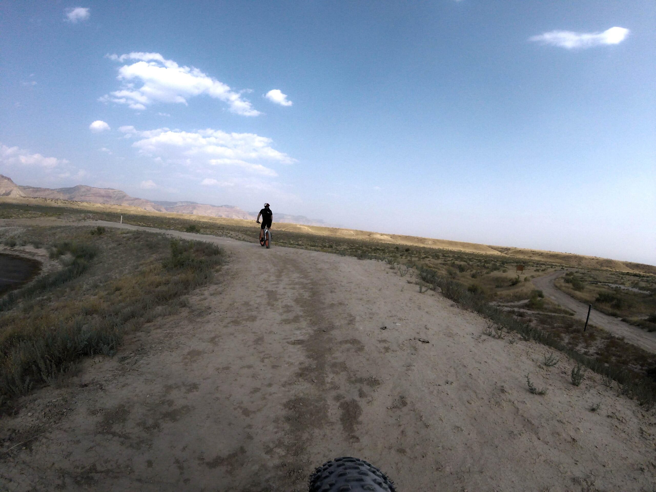 A view of a cyclist riding on a dirt trail that winds through a vast, open landscape with low shrubs and distant mountains under a partially cloudy sky. 18 Road Trails / North Fruita Desert mountain bike trail.