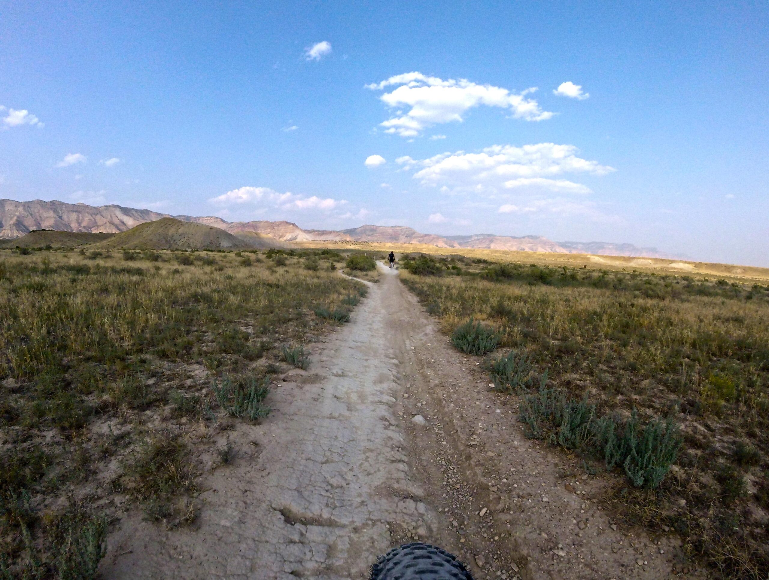A dirt path winding through an open landscape, surrounded by dry grasses and shrubs, with rolling hills and distant mountains under a partly cloudy blue sky. A person can be seen riding a bicycle along the trail. 18 Road Trails / North Fruita Desert mountain bike trail.