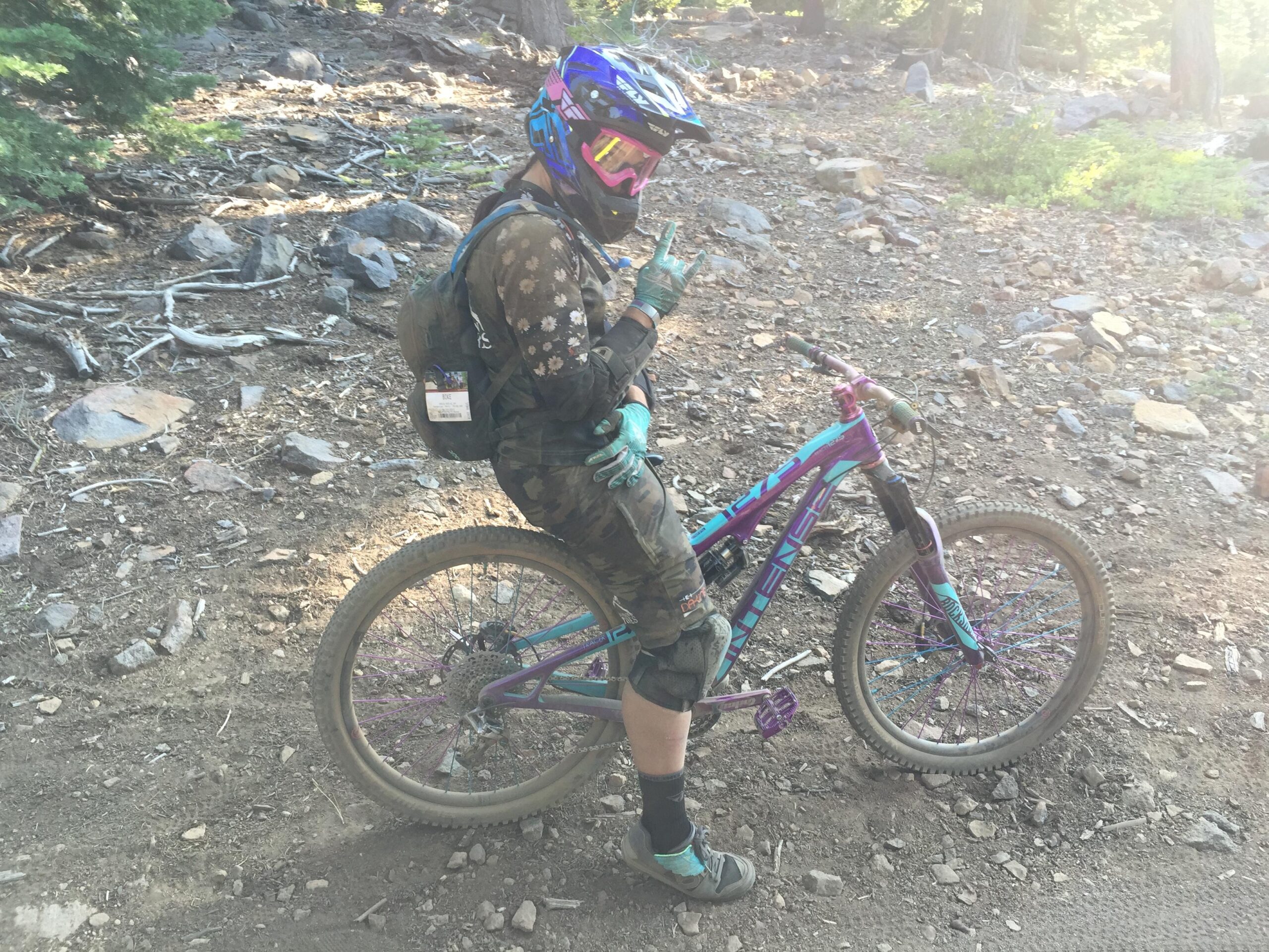 Intense Tracer 275: A person in a camouflaged outfit is sitting on a brightly colored mountain bike on a rocky trail in a forested area. They are wearing a helmet with a visor and gloves, making a hand gesture that resembles a "rock on" sign. The background features trees and scattered rocks.