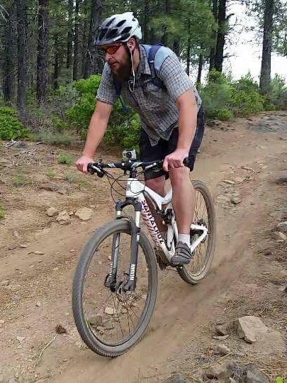 A person riding a mountain bike on a dirt trail surrounded by trees, wearing a helmet and sunglasses. The rider is dressed in a short-sleeved plaid shirt and shorts, focused on navigating the rocky terrain. Phil's Area mountain bike trail.