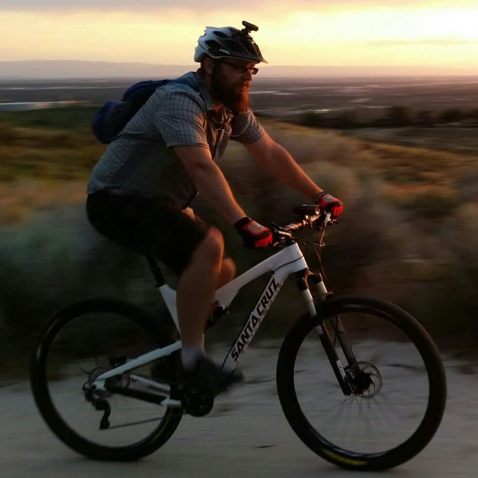 Santa Cruz Tallboy Carbon: A person wearing a helmet and sunglasses rides a white mountain bike along a gravel trail at sunset. The scene captures a dynamic motion, with the bike's wheels slightly blurred to emphasize speed. The background features rolling hills and a colorful sky transitioning from orange to purple. The rider is dressed in a short-sleeve shirt and shorts, with a backpack strapped on their back.