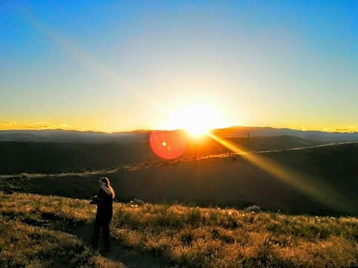 A person standing on a hillside, silhouetted against a vibrant sunset. The sun is just above the horizon, casting warm rays across the landscape and creating lens flares. The sky is a gradient of blue to orange, with rolling hills in the background. Ada/Eagle Bike Park mountain bike trail.