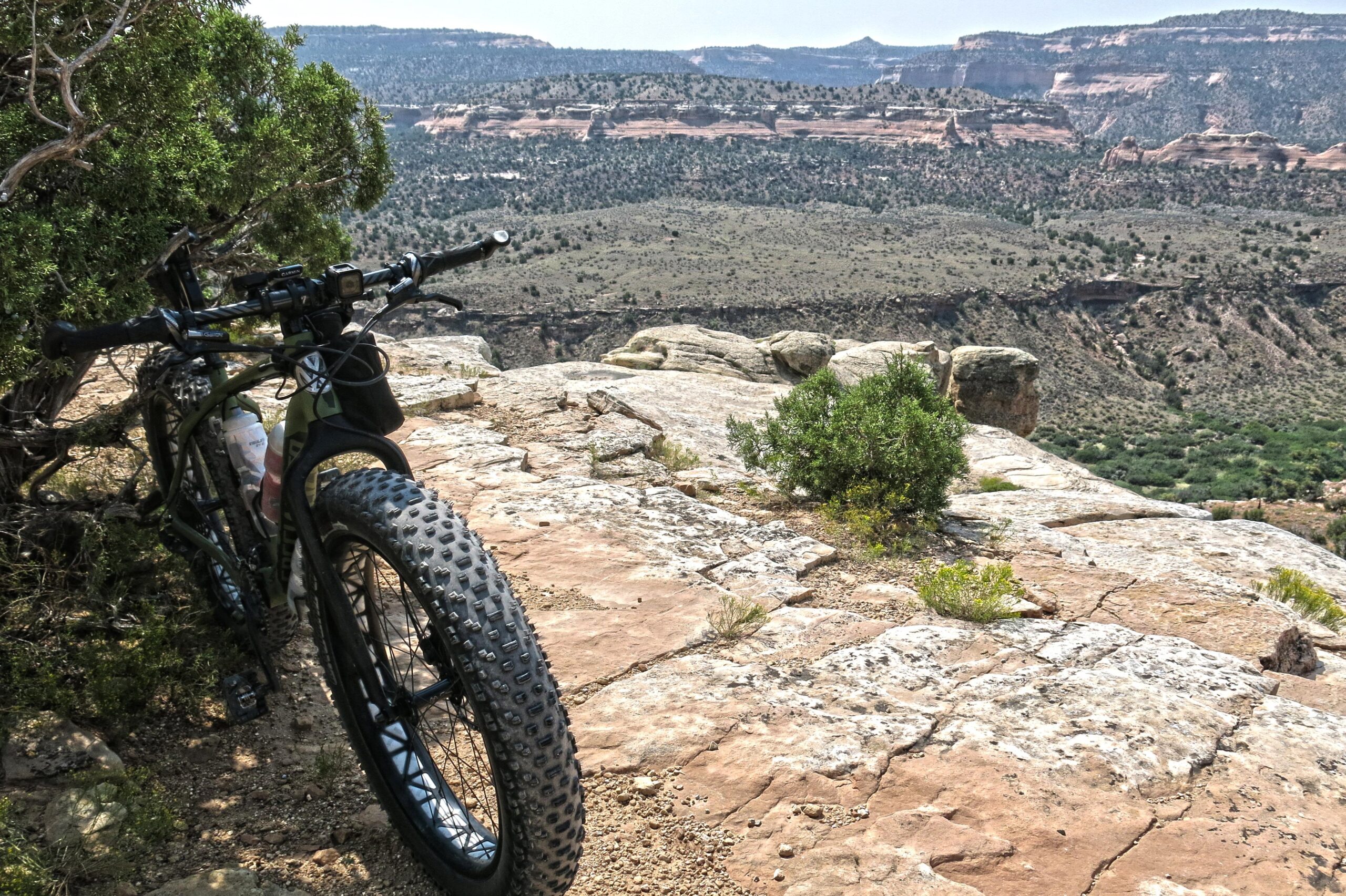 A mountain bike with thick tires rests on a rocky outcrop, overlooking a scenic view of rolling hills and distant cliffs. Sparse vegetation grows on the rocky terrain, framed by expansive, arid landscape. Mary's Loop / Horsethief Bench mountain bike trail.