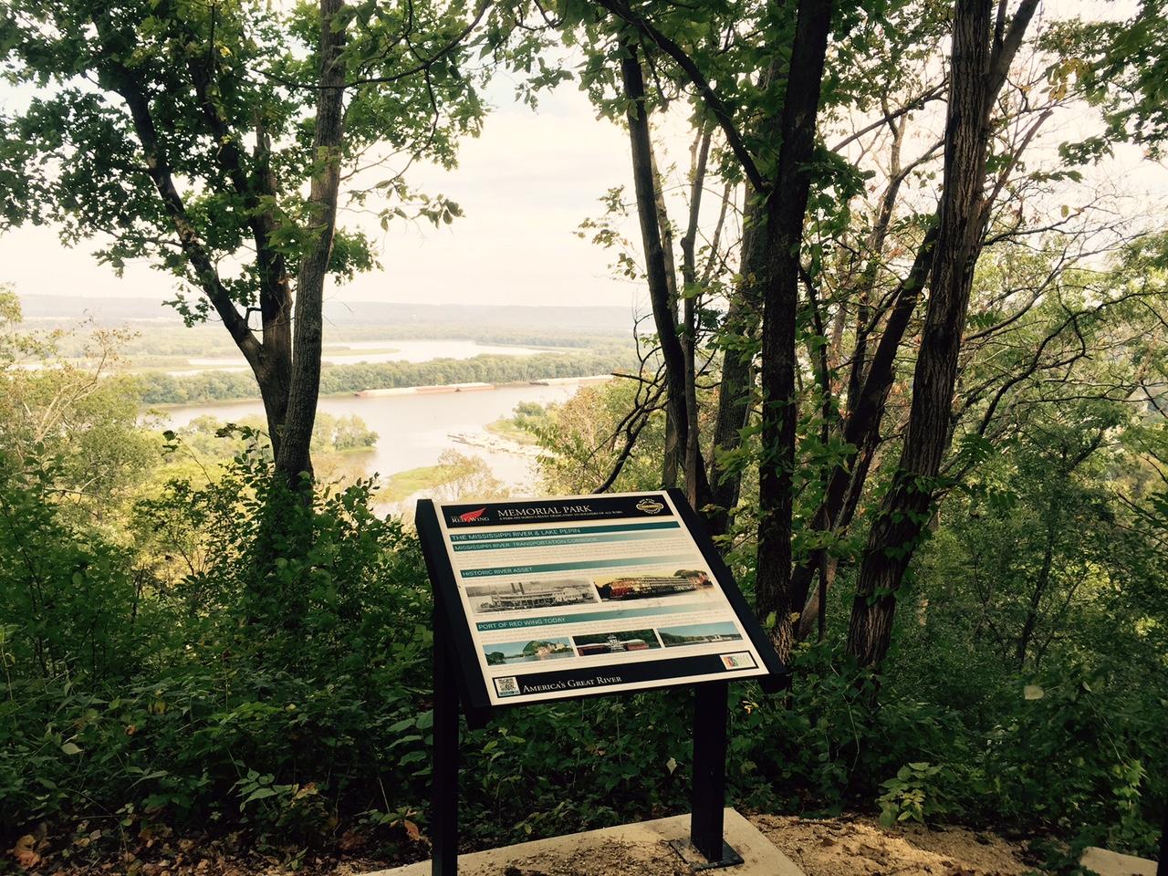 Scenic view from a hillside in a park, featuring a sign that provides information about the area. Lush green trees frame the setting, with a river winding through the landscape below. The sky is partly cloudy, enhancing the natural beauty of the scene. Memorial Park mountain bike trail.
