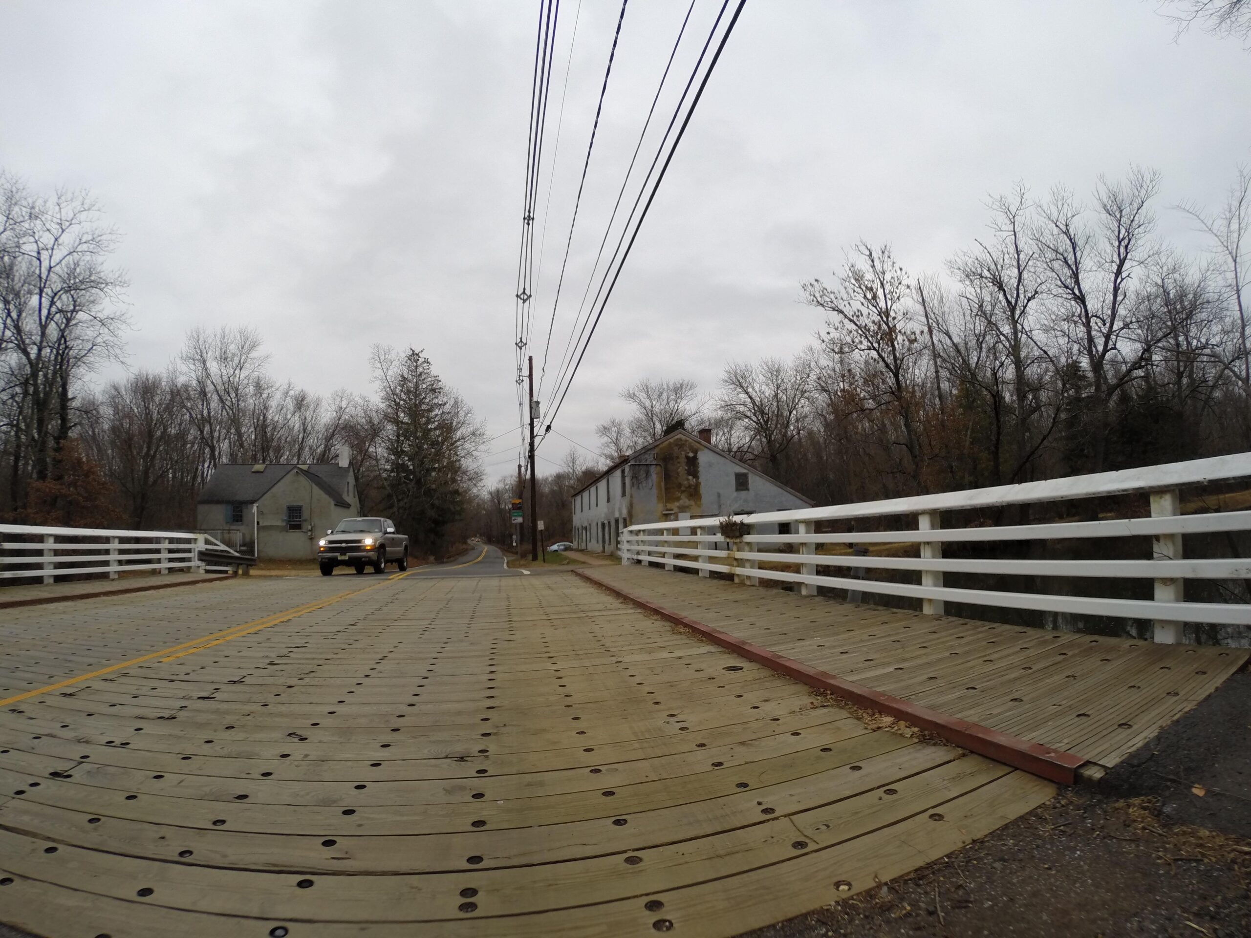 A wooden bridge spans a road lined with power lines, with a pickup truck driving towards the camera. On the left, a gray house is partially visible, surrounded by bare trees and a cloudy sky. In the background, a weathered building can be seen along the roadside. Bridge To Bridge - D&r Canal mountain bike trail.