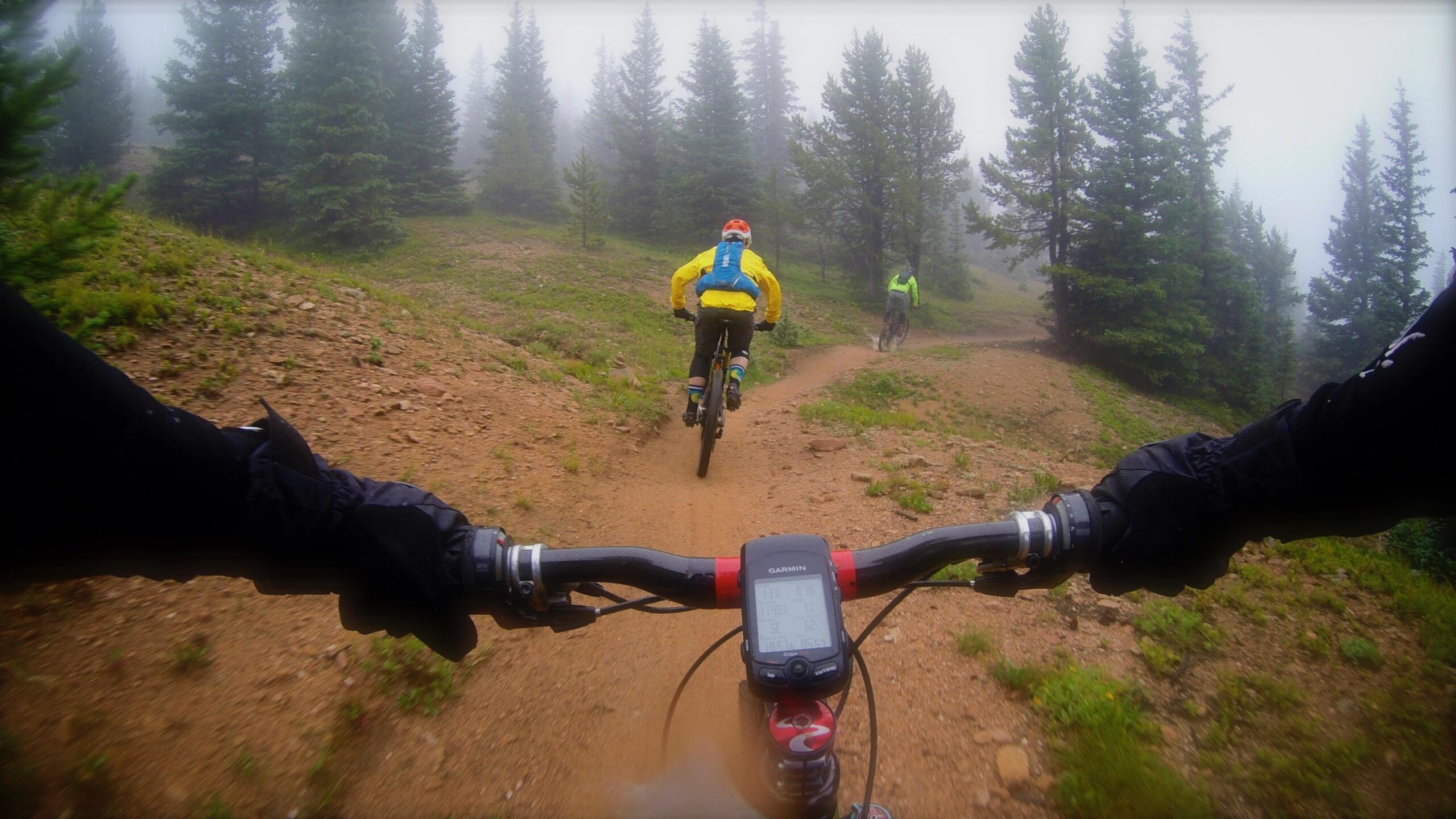 A close-up view of a mountain bike handlebar in a misty forest, with two cyclists riding along a dirt trail ahead. The foreground shows the bike's display, while the background features evergreen trees and a foggy atmosphere. One rider wears a bright yellow jacket, and the other is dressed in green, emphasizing the outdoor adventure theme. Monarch Crest Trail mountain bike trail.