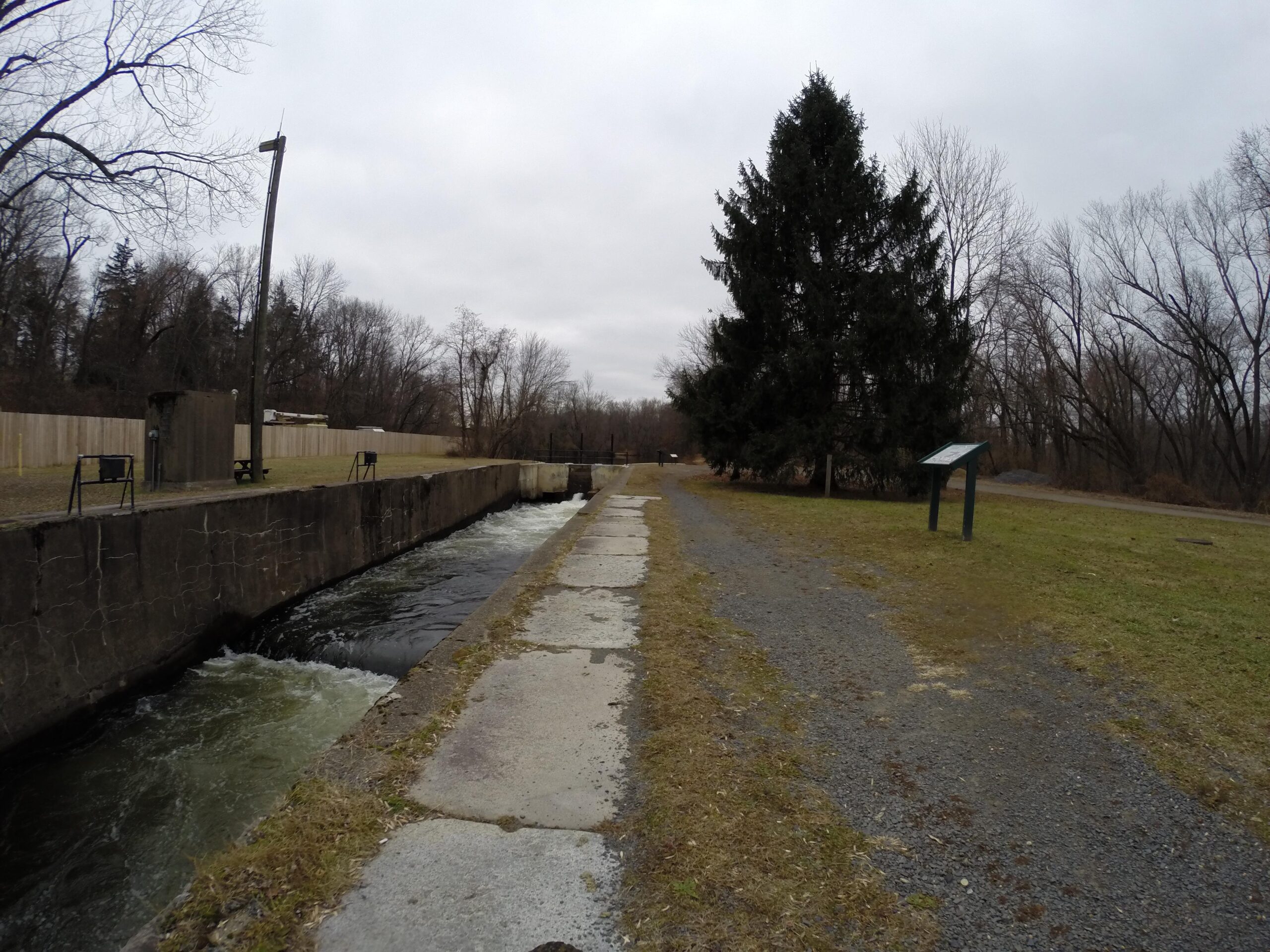 Landscape view of a canal with flowing water, bordered by a stone wall and a grassy path. A tall tree is visible on the right, and an informational sign stands nearby. The scene is set on a cloudy day, reflecting a tranquil outdoor environment with minimal human presence. Bridge To Bridge - D&r Canal mountain bike trail.