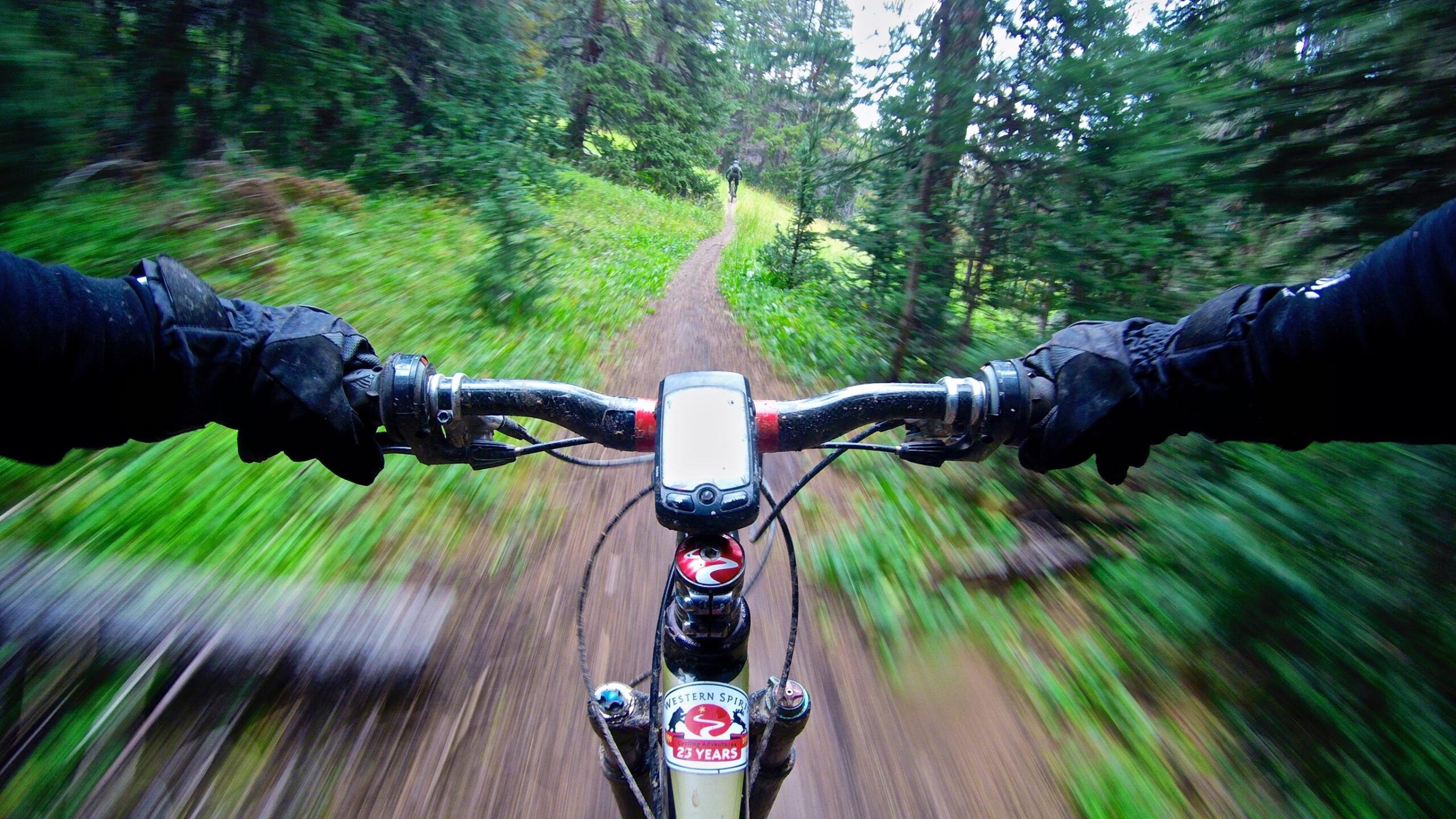Mountain bike handlebars seen from a rider's perspective, with blurred motion indicating speed. Lush green foliage and a dirt trail are visible in the background, suggesting a forested outdoor setting. The handlebars are equipped with a cycling computer and a sticker on the bike frame celebrating 23 years of a biking program. Monarch Crest Trail mountain bike trail.