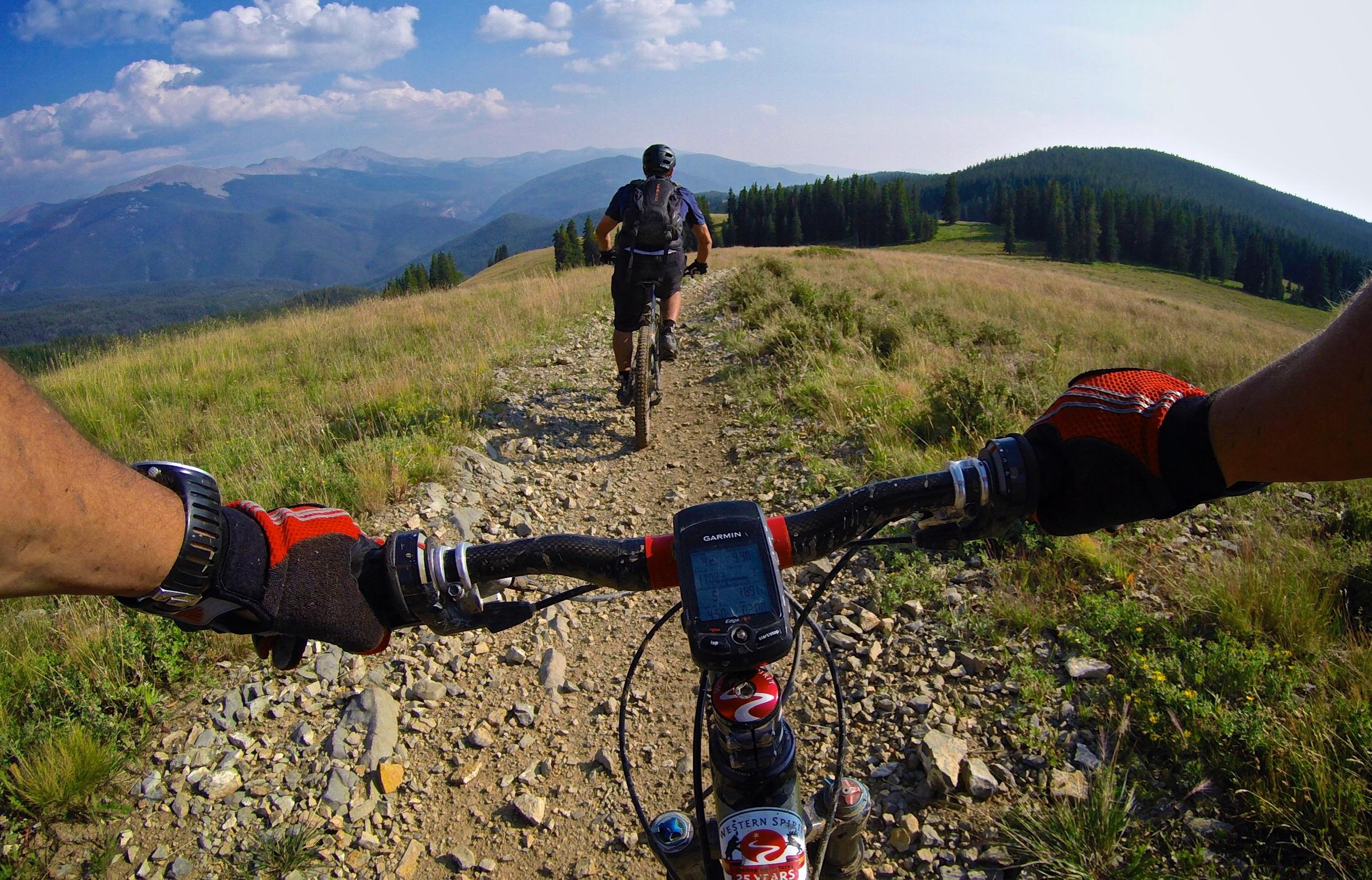 Two mountain bikers navigating a rocky trail through a grassy hillside, with a scenic view of mountains and blue sky in the background. The handlebars of one bike and a GPS device are visible in the foreground, showcasing the rugged terrain. Doctor Park mountain bike trail.