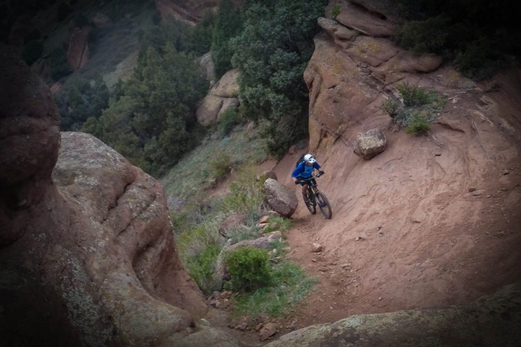 A mountain biker in a blue jacket navigates a rocky, sloped trail surrounded by greenery and rugged terrain. The image captures the cyclist mid-motion as they descend along the path, showcasing the challenging outdoor environment.