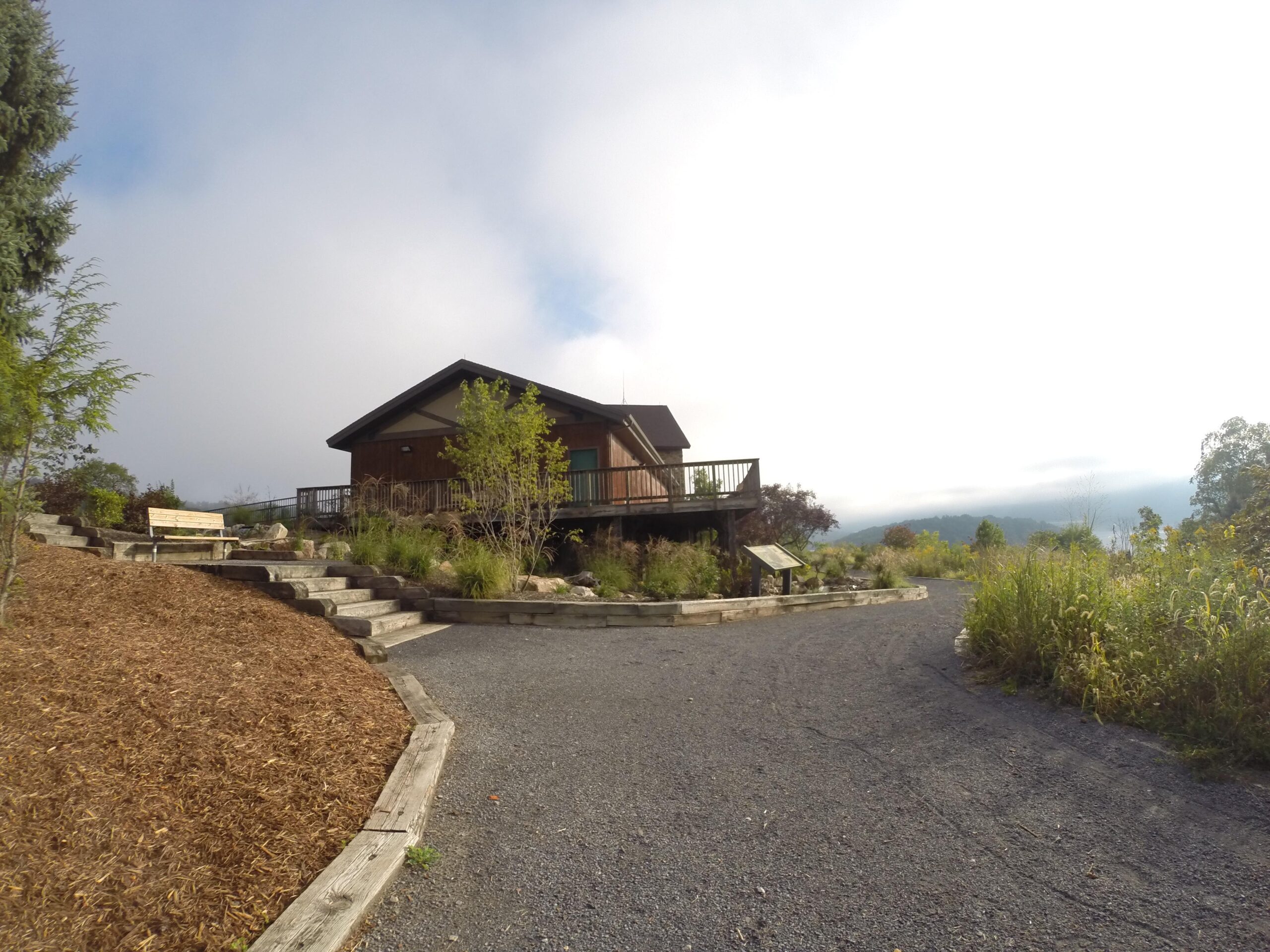A wooden house with a deck sits on a hillside surrounded by greenery and wildflowers. A stone pathway leads up to the house, with a wooden bench nearby. The sky is cloudy, creating a serene atmosphere. Allegrippis Trails mountain bike trail.