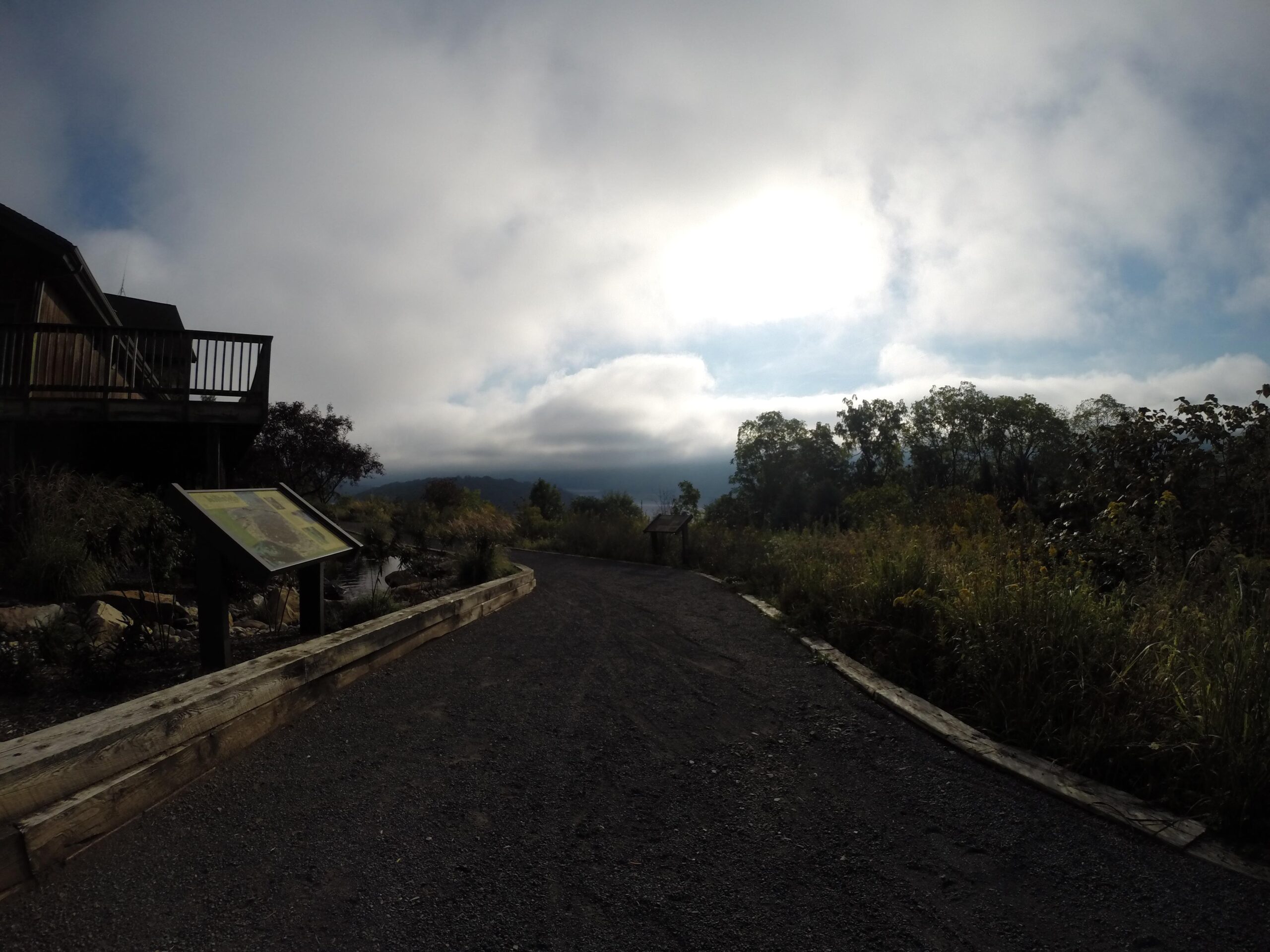 A gravel path leads through a natural setting, with tall grasses and plants on either side. In the foreground, there is a wooden sign with informational content. To the left, a building with a balcony is partially visible, and the sky above is partly cloudy with hints of sunlight peeking through. Trees are seen in the distance, creating a serene outdoor atmosphere. Allegrippis Trails mountain bike trail.