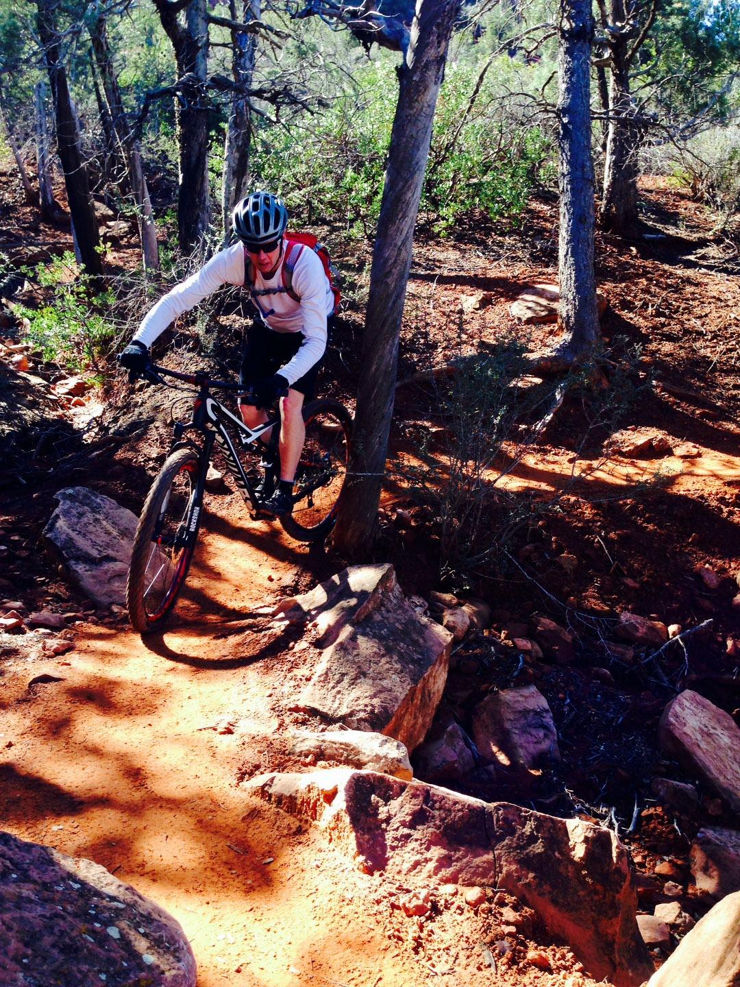 Specialized Camber SW 29: A mountain biker navigating a rocky trail through a forested area, with sunlight filtering through the trees. The cyclist is wearing a helmet and gloves, focused on maneuvering around boulders and rough terrain. The path is lined with reddish dirt and small shrubs.
