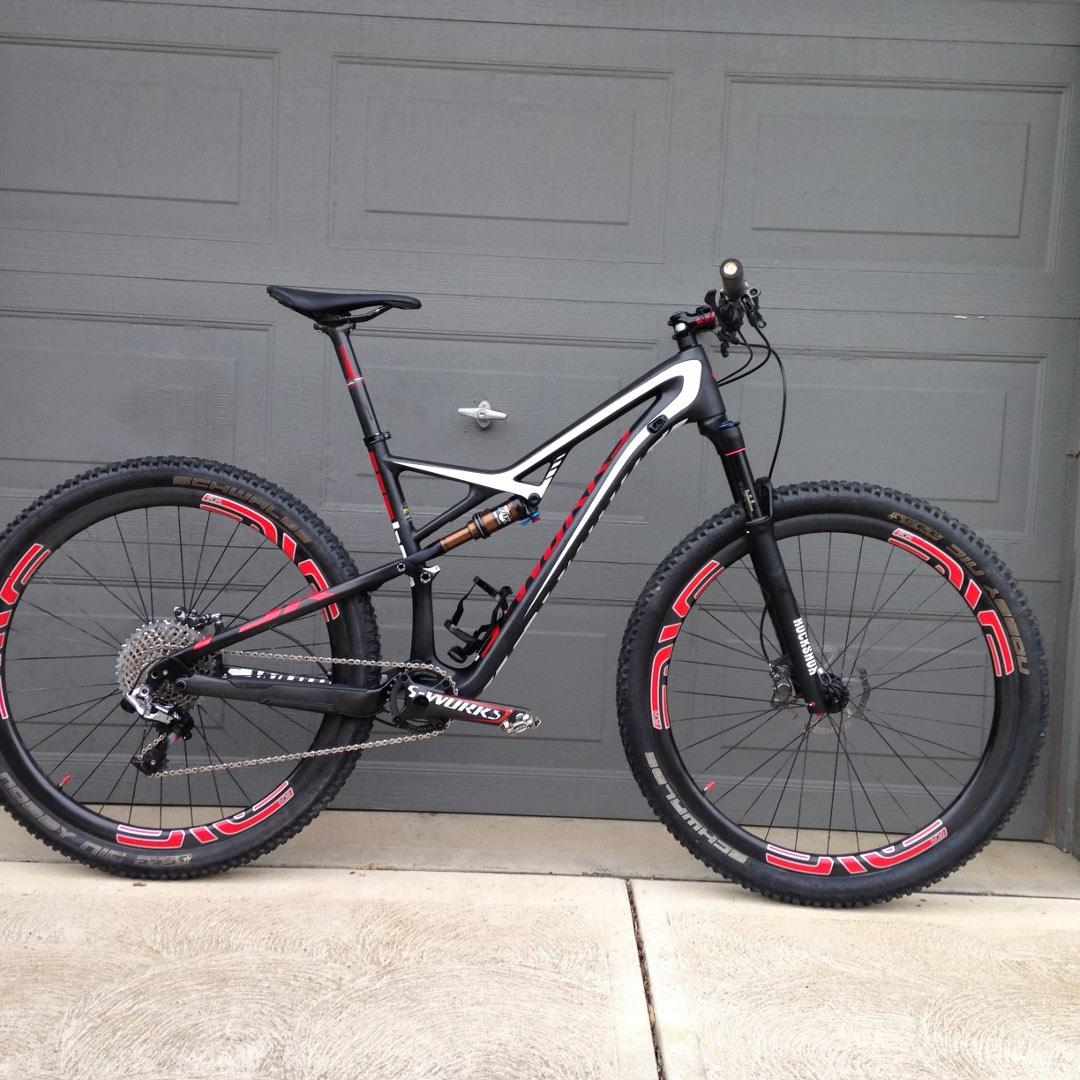 Specialized Camber SW 29: A black and white mountain bike with red accents, featuring large knobby tires, a sleek frame, and a suspension system, positioned against a gray garage door.