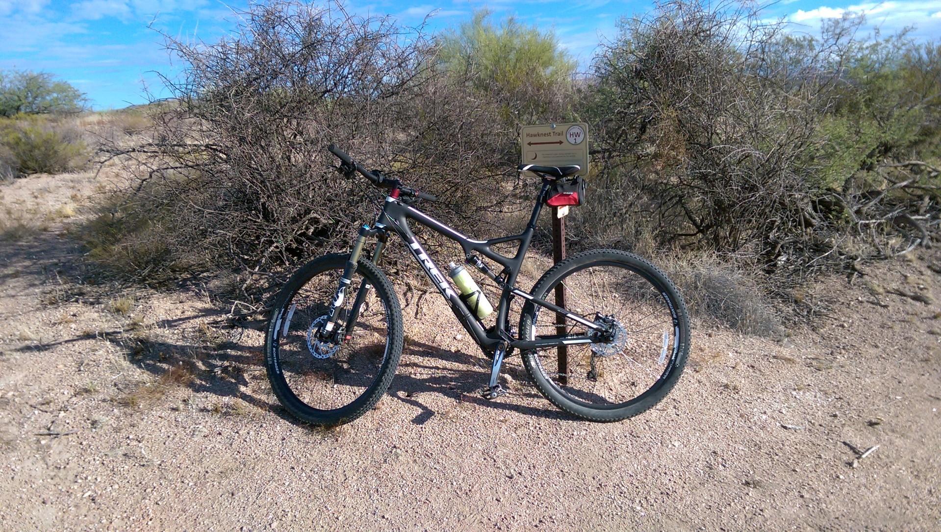 A mountain bike parked on a gravel trail surrounded by sparse desert vegetation, with a trail signpost labeled "Hawknest Trail" in the background under a clear blue sky. Brown's Ranch to Granite Mountain mountain bike trail.