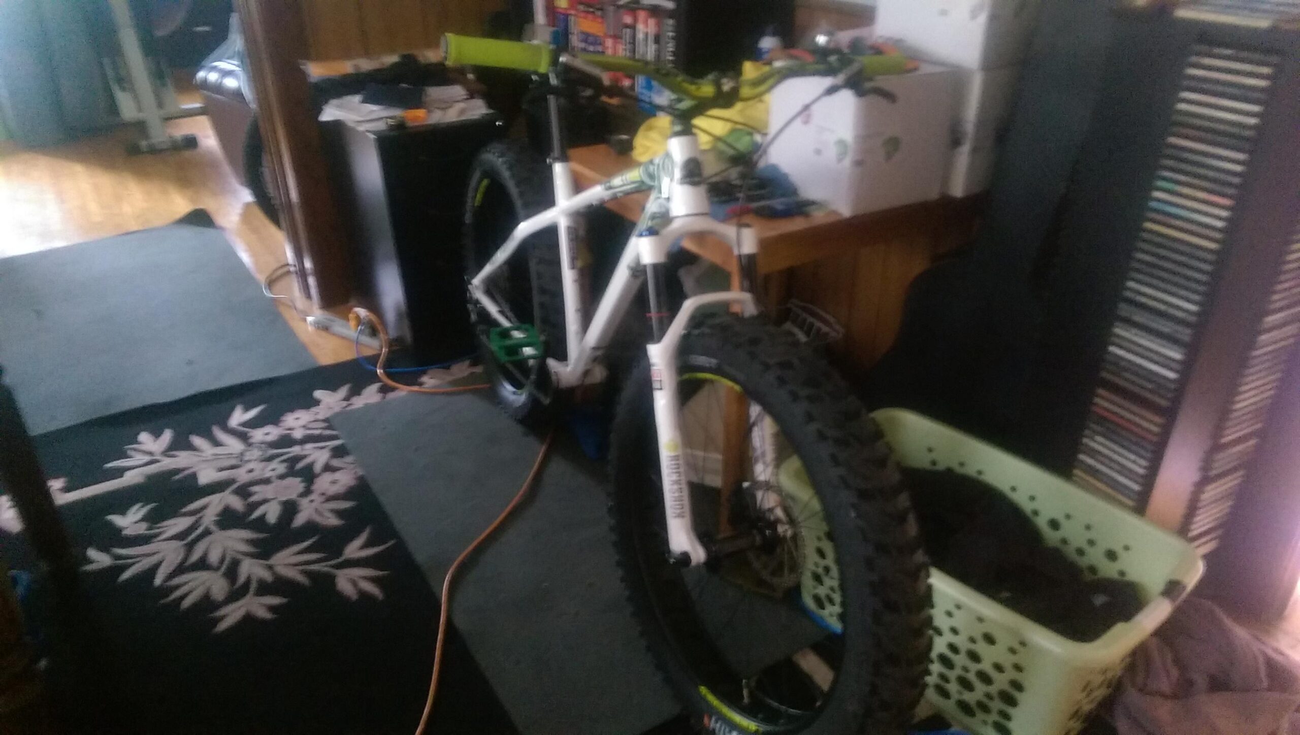 A white fat bike with green handlebars and large tires is positioned in a cluttered room. In the background, various items can be seen, including a wooden table, a basket of laundry, and a shelf filled with CDs. The flooring features a patterned rug, and there are cables on the floor.