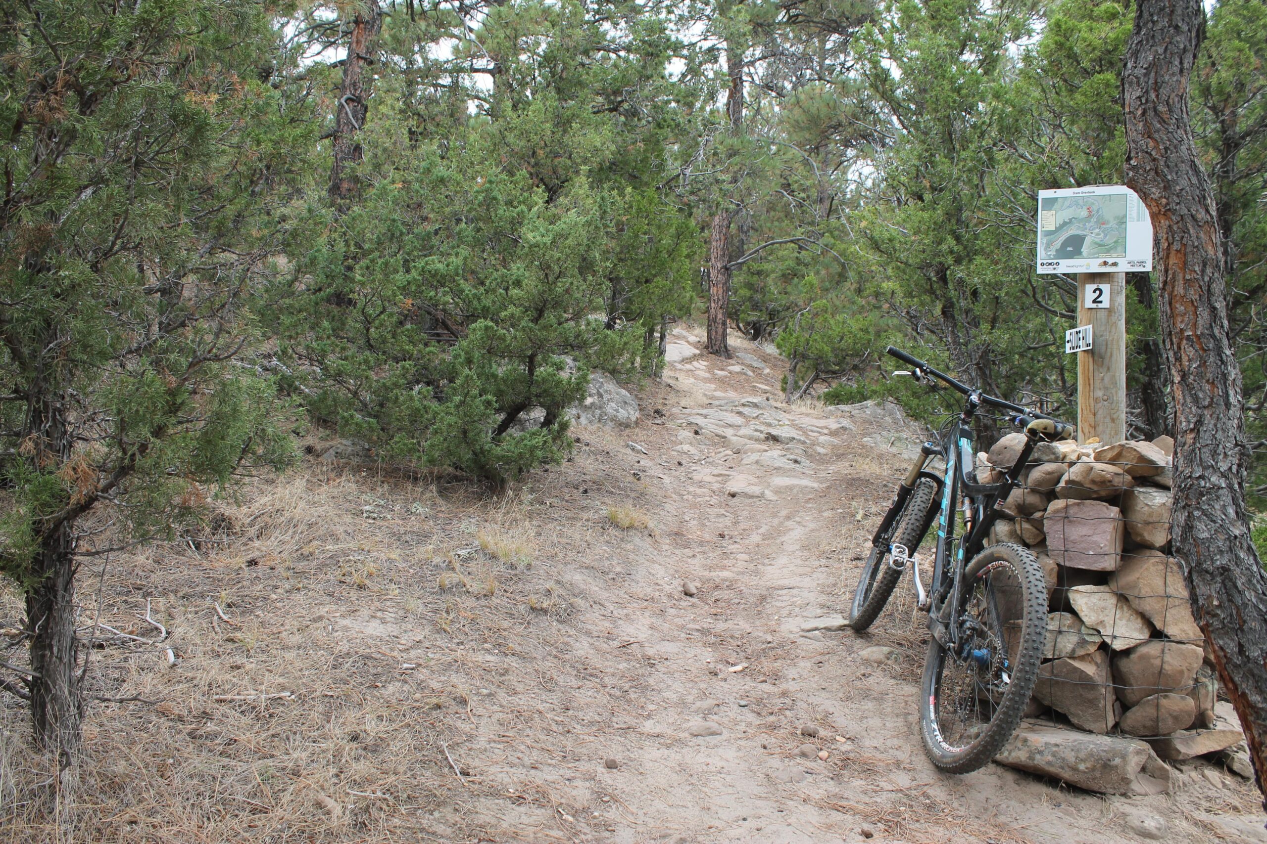 A mountain bike leaned against a stone marker along a narrow trail surrounded by trees. The path is rocky and dusty, indicating a natural outdoor setting. A sign with a map is visible in the background, positioned on a post next to the trail. Glendo State Park mountain bike trail.
