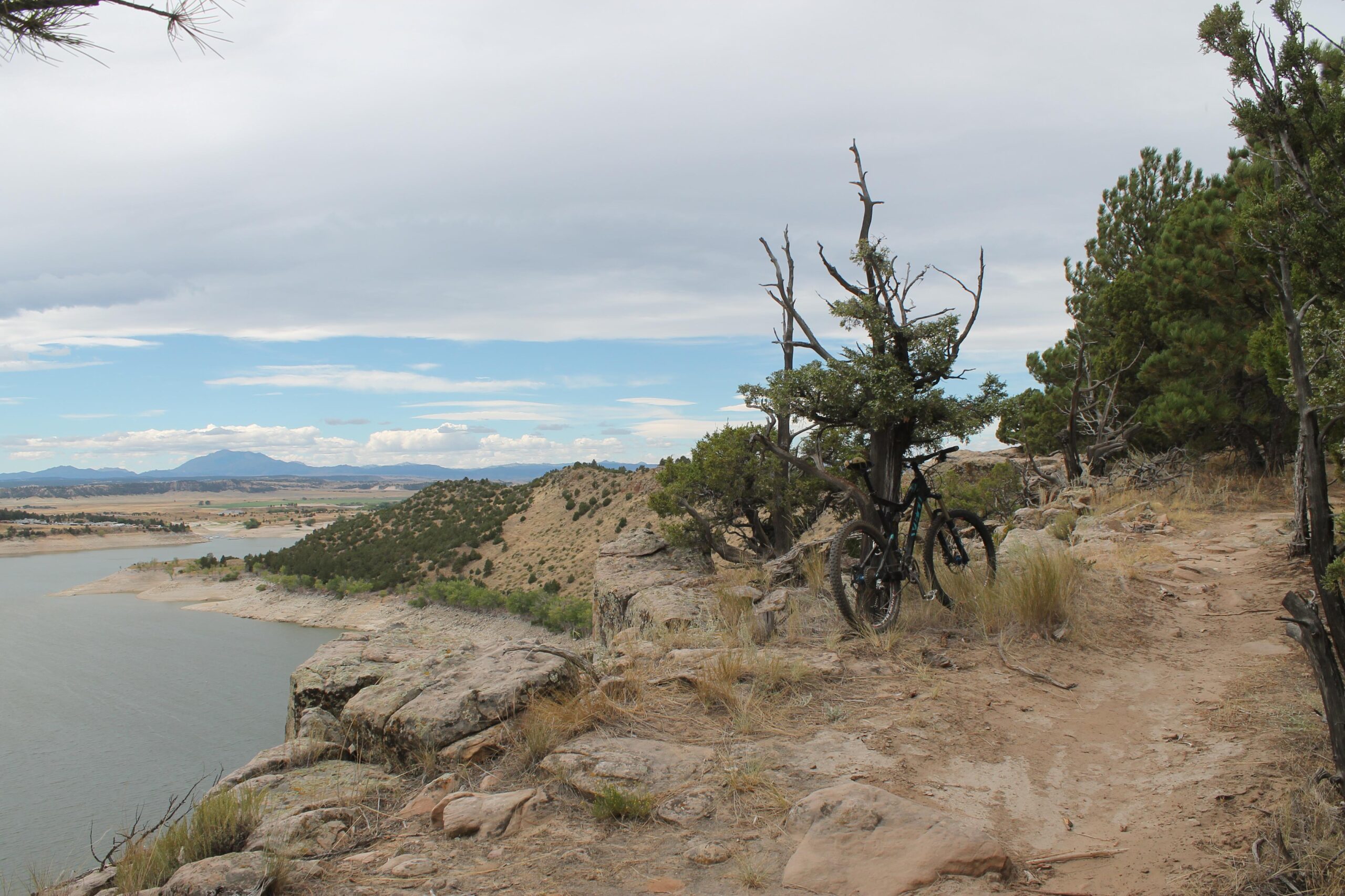 Mountain biking trail overlooking a lake, surrounded by rocky terrain and pine trees, under a cloudy sky. A mountain bike is resting against a bush near the trail, with hills and distant mountains visible in the background. Glendo State Park mountain bike trail.