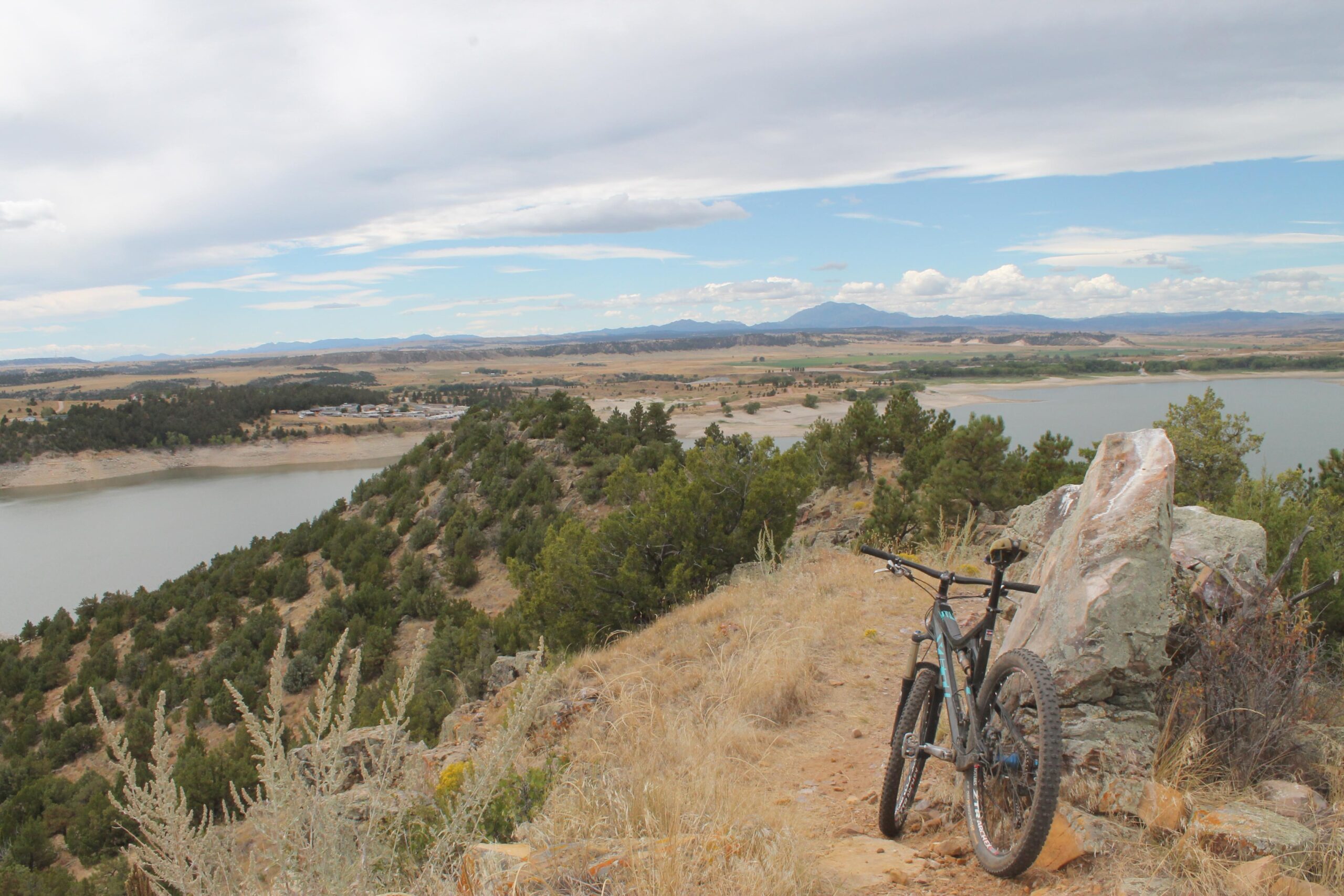 A mountain bike leaning against a large rock on a hillside, overlooking a scenic view of a lake and distant mountains. The landscape features rolling hills, patches of greenery, and a partly cloudy sky. Glendo State Park mountain bike trail.