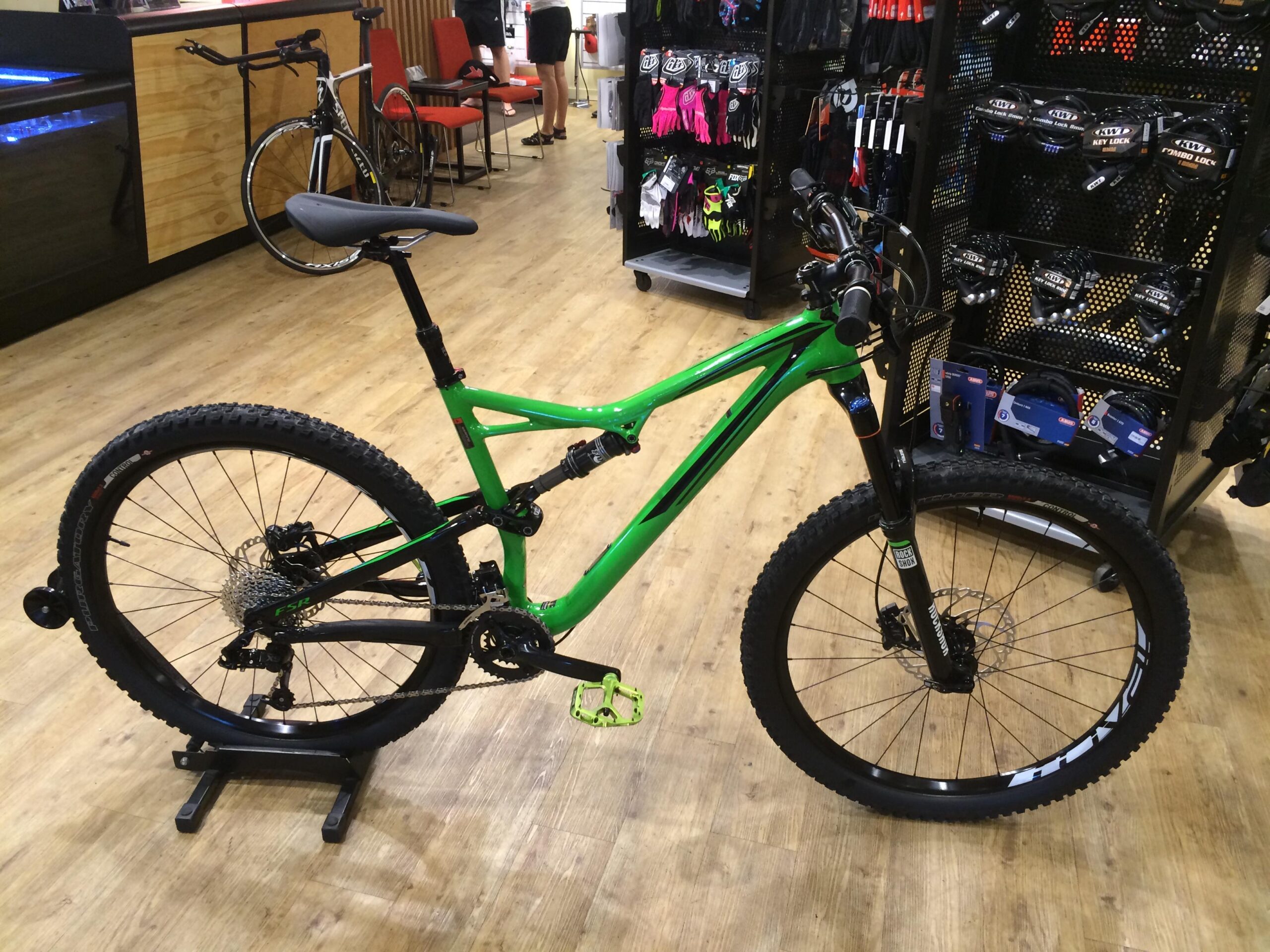 Specialized Stumpjumper FSR: A green mountain bike displayed in a bike shop, featuring a sturdy frame, thick tires, and a front suspension fork. The bike is positioned on a stand, with various biking accessories and gear visible in the background, including gloves and locks. The shop has a wooden floor and casual seating in the vicinity.