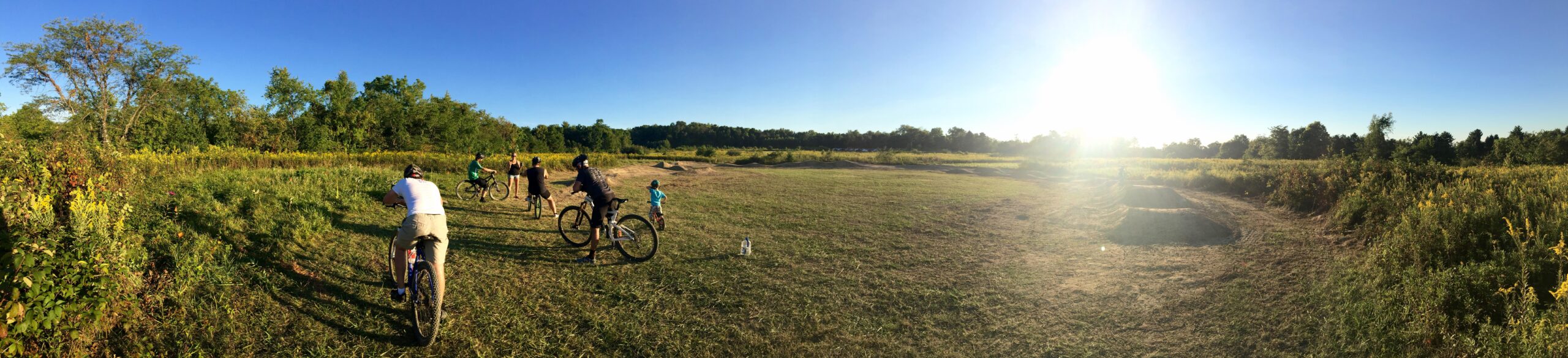 A panoramic view of a grassy field during golden hour, featuring several people on bicycles. In the foreground, two cyclists are positioned facing away from the camera, while others gather nearby. The landscape is dotted with trees and greenery, with a bright sun setting in the background, casting a warm light over the scene. Dirt bike jump ramps are visible in the distance, suggesting an active recreational area. Chestnut Ridge mountain bike trail.