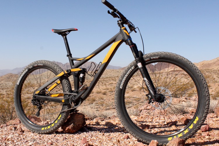 A modern mountain bike with a black and orange frame, positioned on rocky terrain in a desert landscape. The bike features thick tires and a suspension system, with a clear blue sky in the background and mountains visible in the distance.