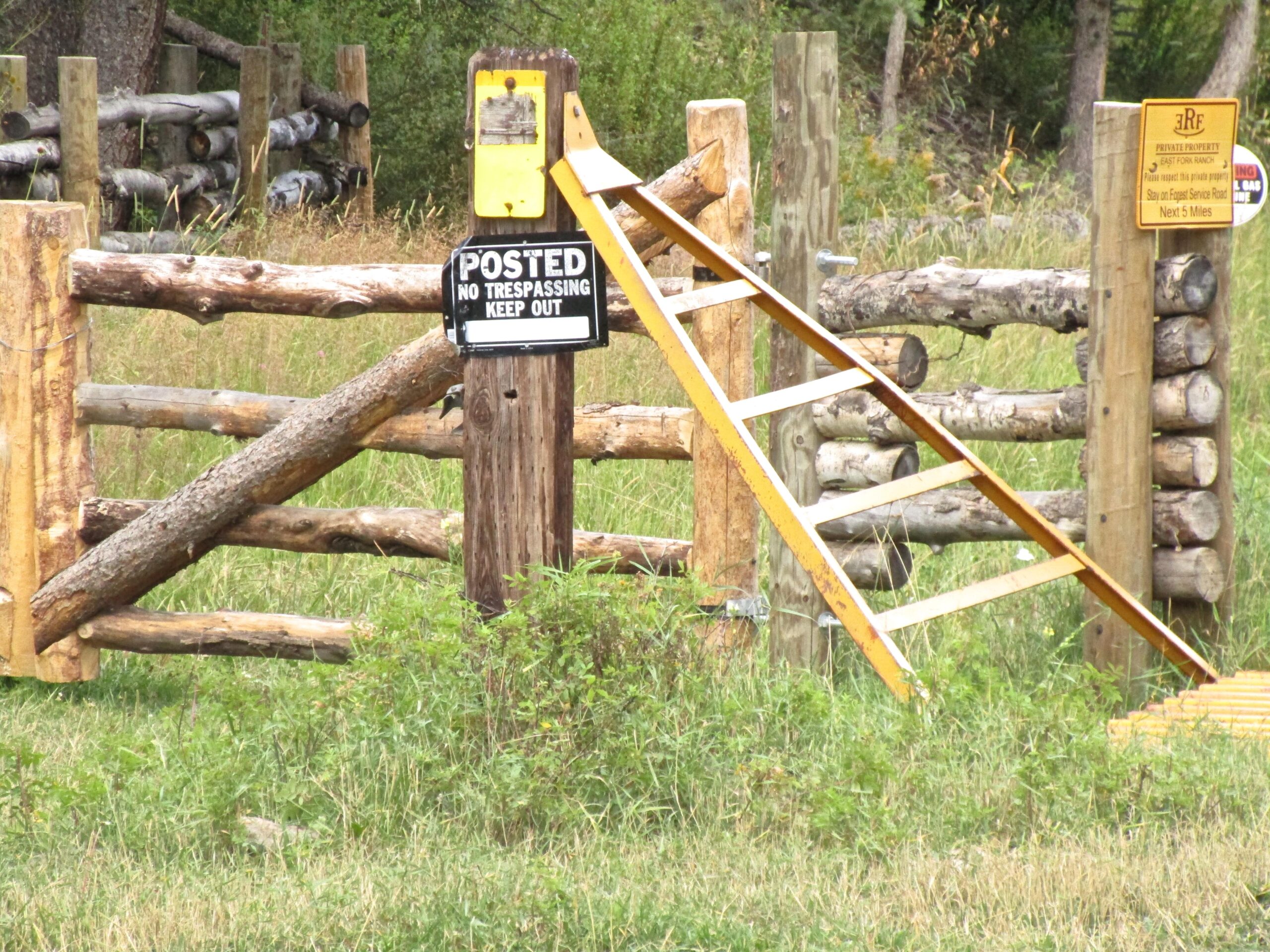A wooden fence with a yellow "Posted No Trespassing Keep Out" sign mounted on a post. A ladder leans against the fence, leading up to a higher section. Adjacent to the sign, another sign indicates "Private Property." The area is grassy and surrounded by trees in the background. Treasure Mountain Trail mountain bike trail.