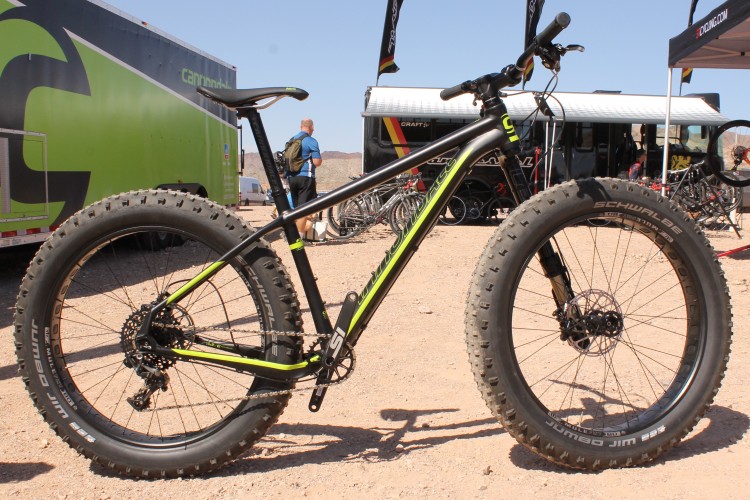 A fat tire mountain bike with a black and neon green frame is positioned in a desert setting. The bike features wide tires designed for off-road terrain, and is parked near a mobile bike display. In the background, there are other bicycles and people, with a focus on outdoor activities. The sun is shining in a clear blue sky.