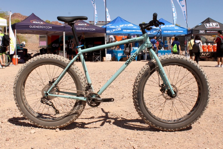 A turquoise Surly bicycle with wide tires is parked on a gravel surface, with various tents and banners in the background promoting energy products. The scene is set in a sunny outdoor environment, suggesting a cycling event or festival.