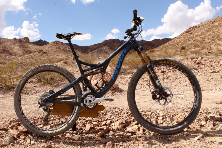 A mountain bike resting on rocky terrain, with a backdrop of rugged hills and a blue sky dotted with clouds. The bike features a black frame with blue accents and is equipped for off-road cycling.